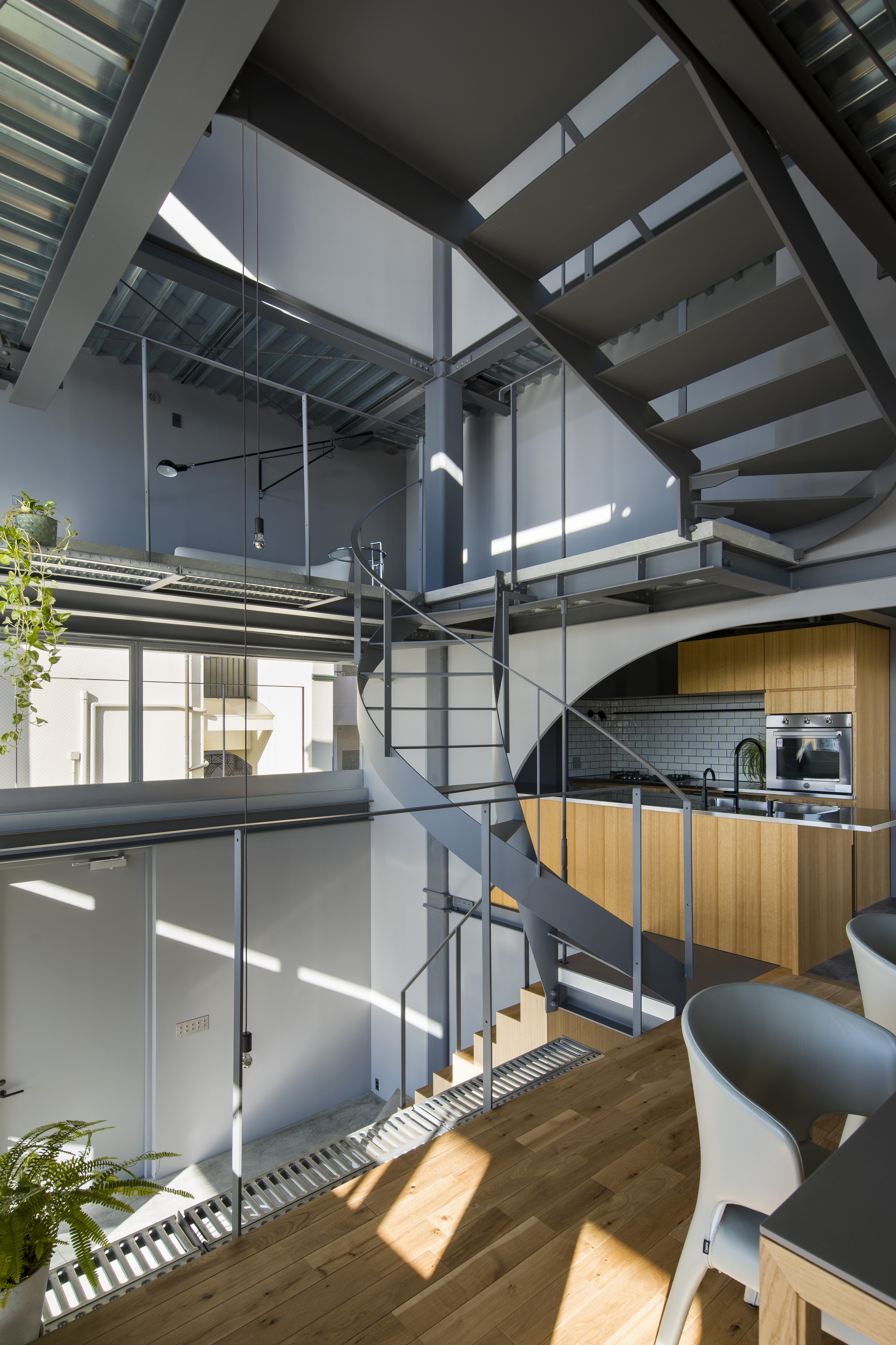 Central spiral stair winding through three floors with timber kitchen cabinetry and steel catwalks illuminated by shafts of sunlight