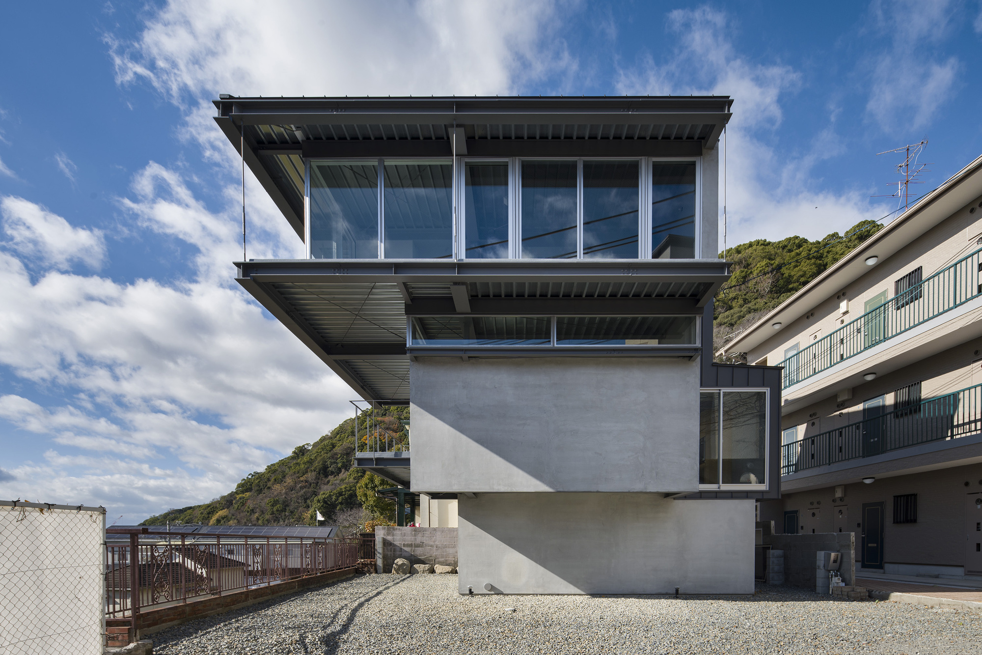 Stacked concrete volumes with cantilevered glass upper floor and gravel courtyard under a partly cloudy sky