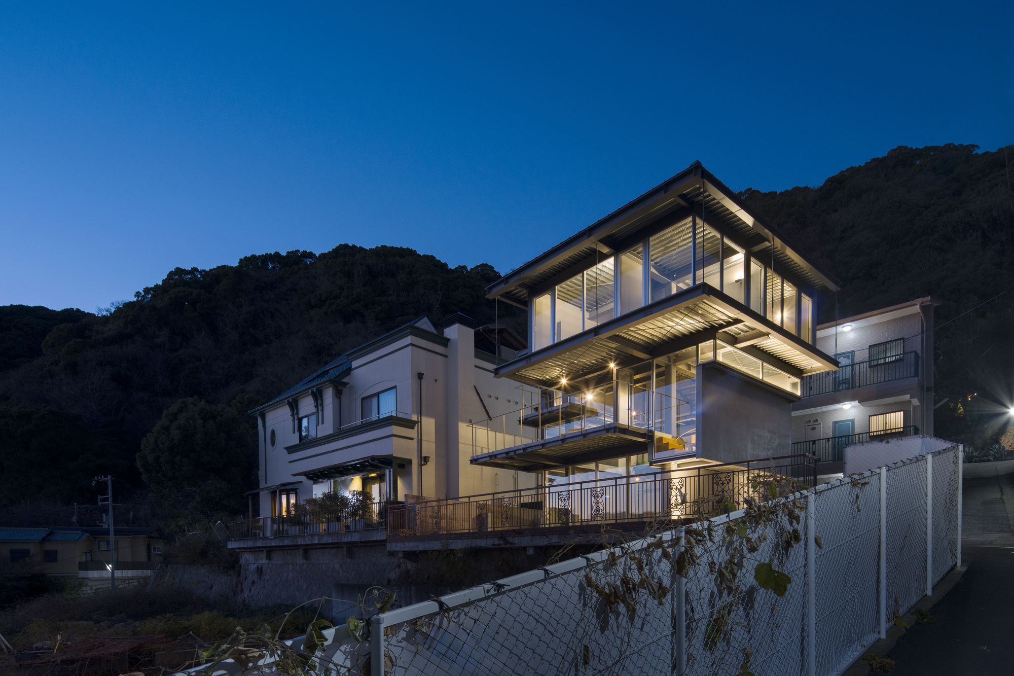 Illuminated steel-framed volume rising above adjacent houses at dusk with forested hillside behind