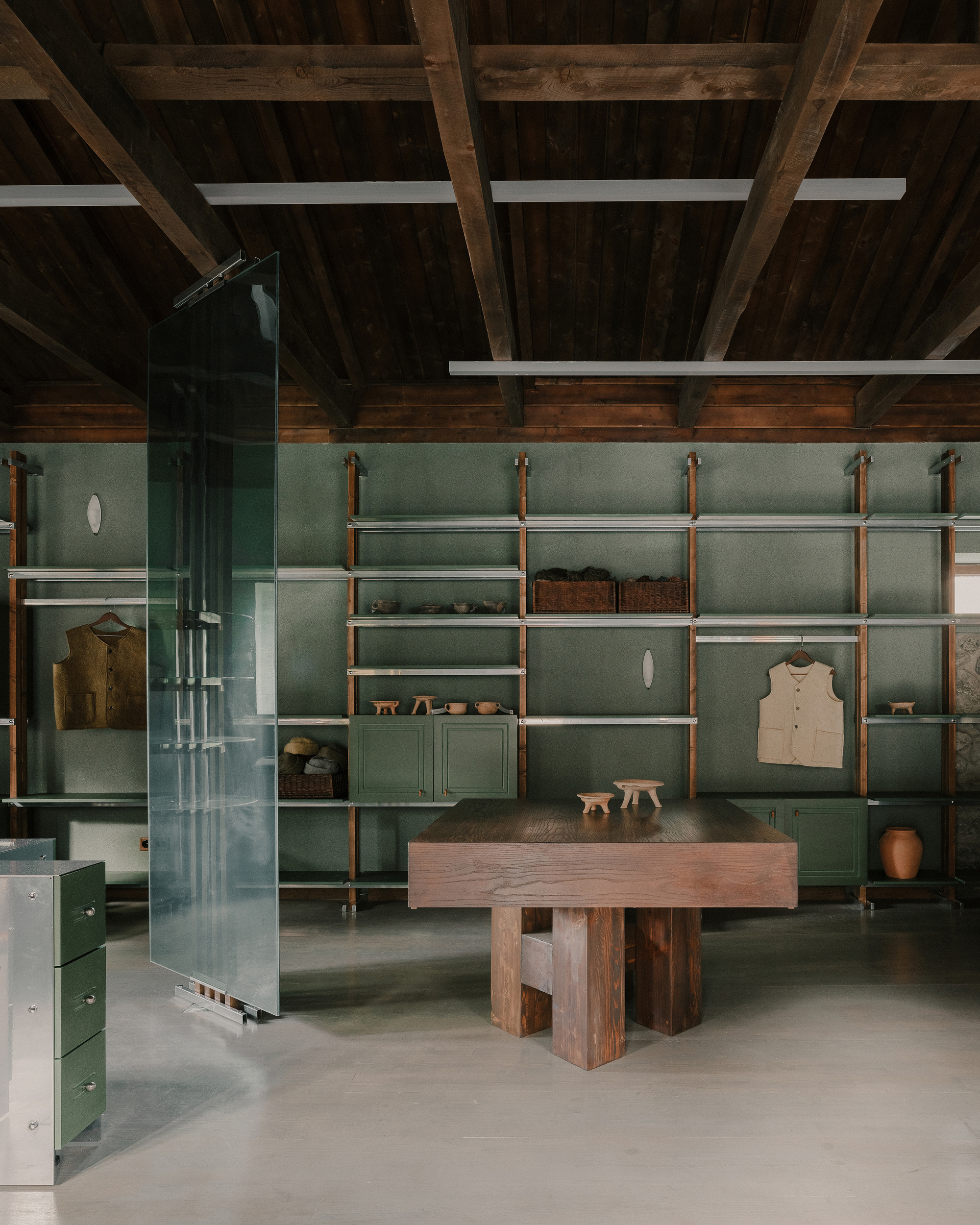 Sage green wall-mounted shelving with timber and metal supports above a solid timber table and polished concrete floor