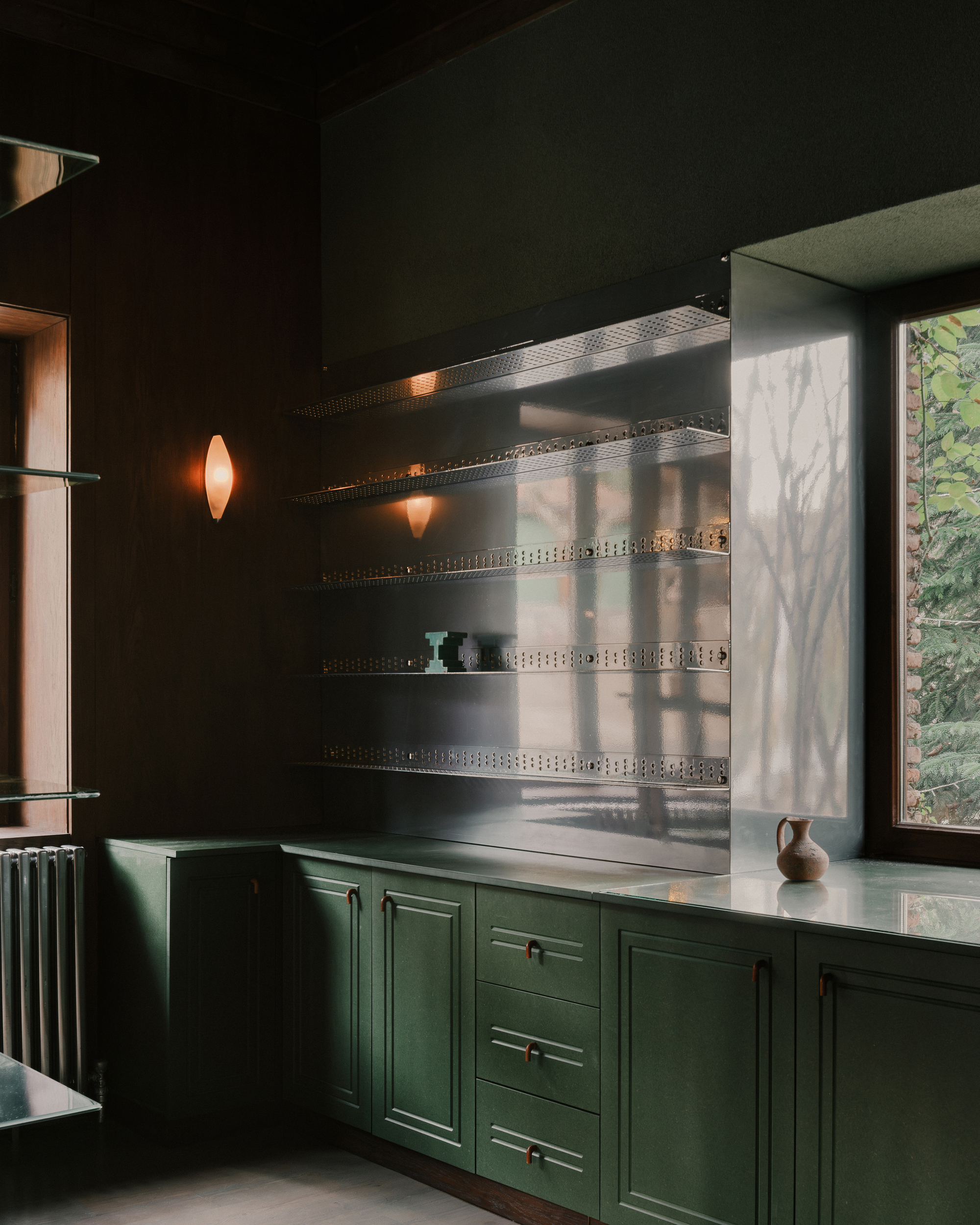 Kitchen alcove with green cabinetry, perforated metal shelves, and sunlight filtering through a window with foliage outside