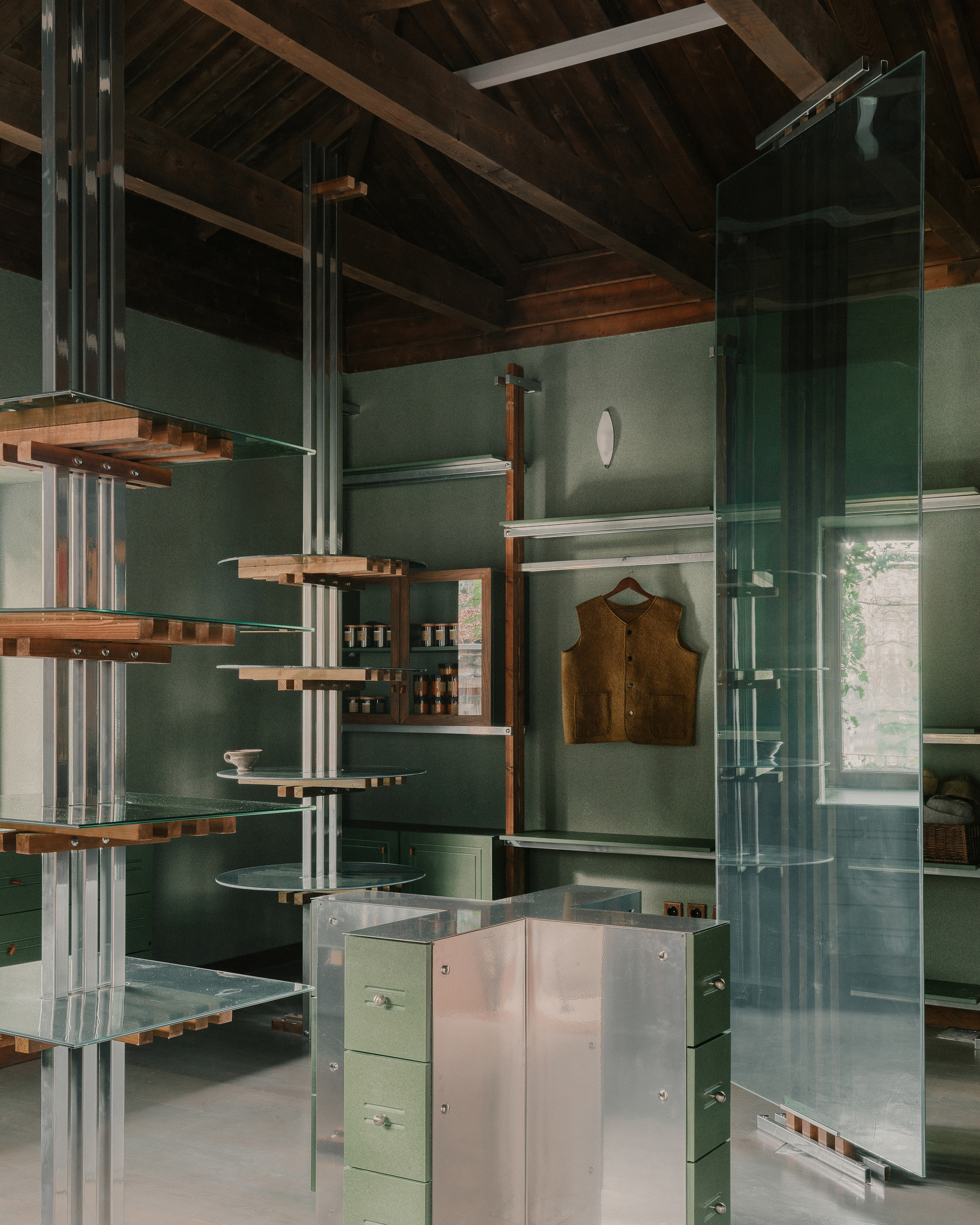 Vertical shelving system with glass shelves suspended between steel tubes against a green paneled wall