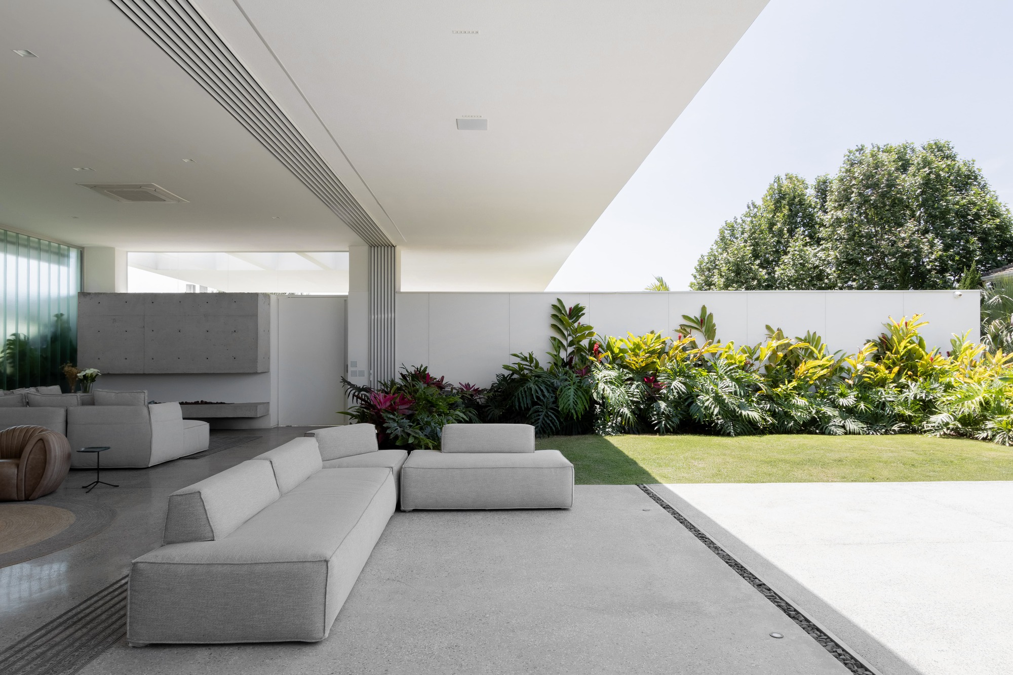 Open-air living area with white sofas on concrete flooring beside a planted garden bed