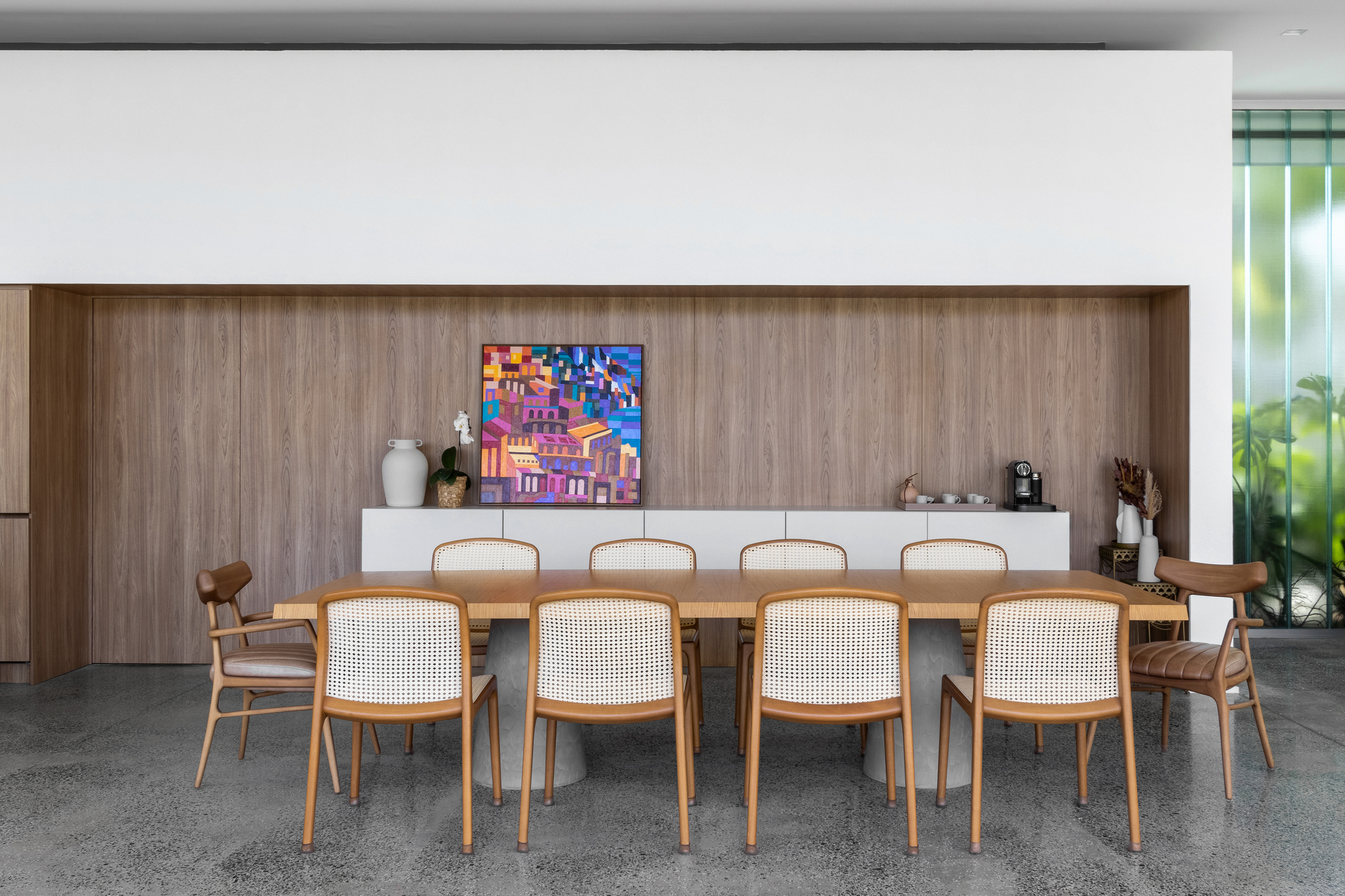 Dining table with cane-backed chairs facing a walnut-paneled wall with low white credenza and colorful artwork