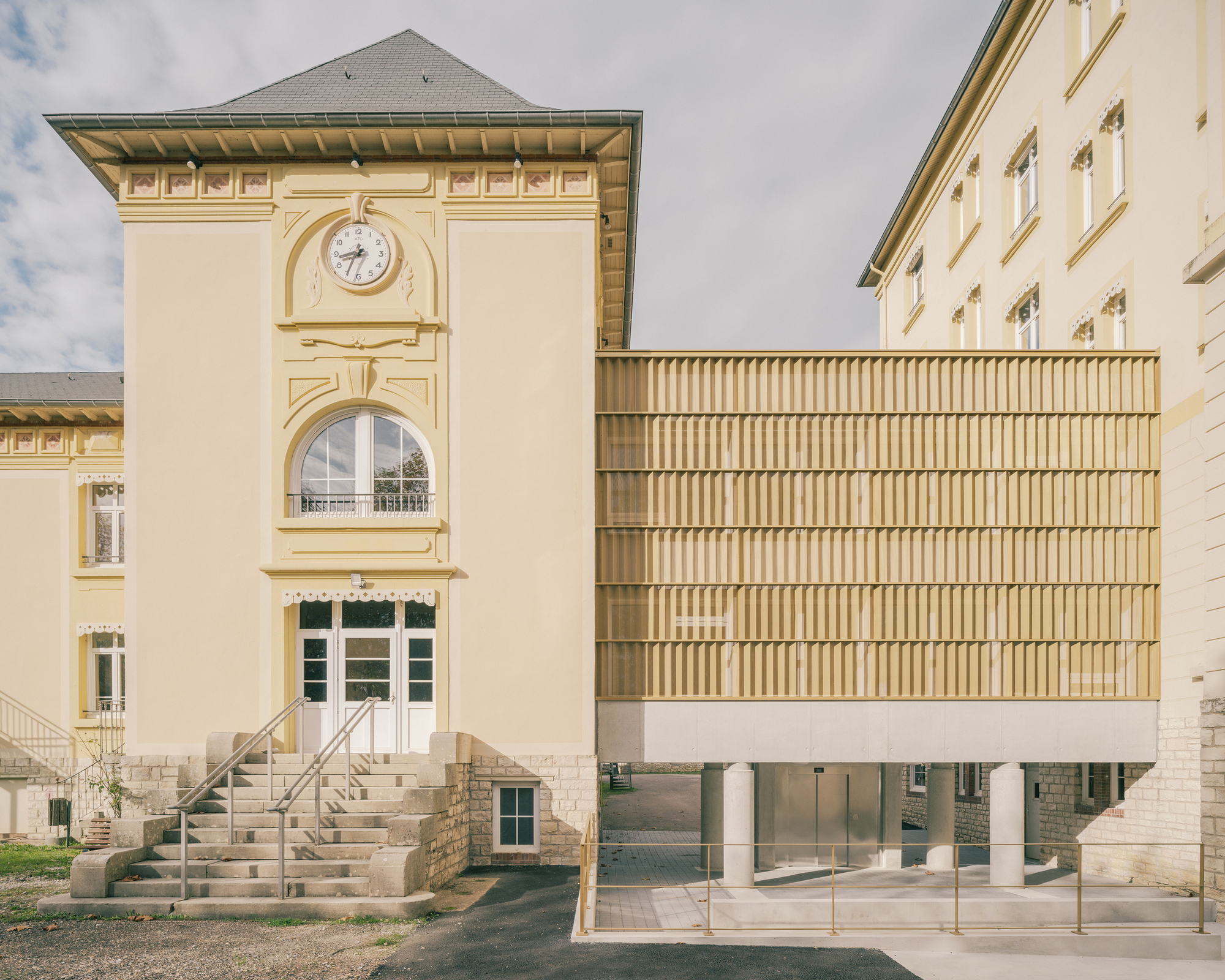 Vertical louvered timber facade on pilotis juxtaposed with a pale yellow clock tower and stone stairs