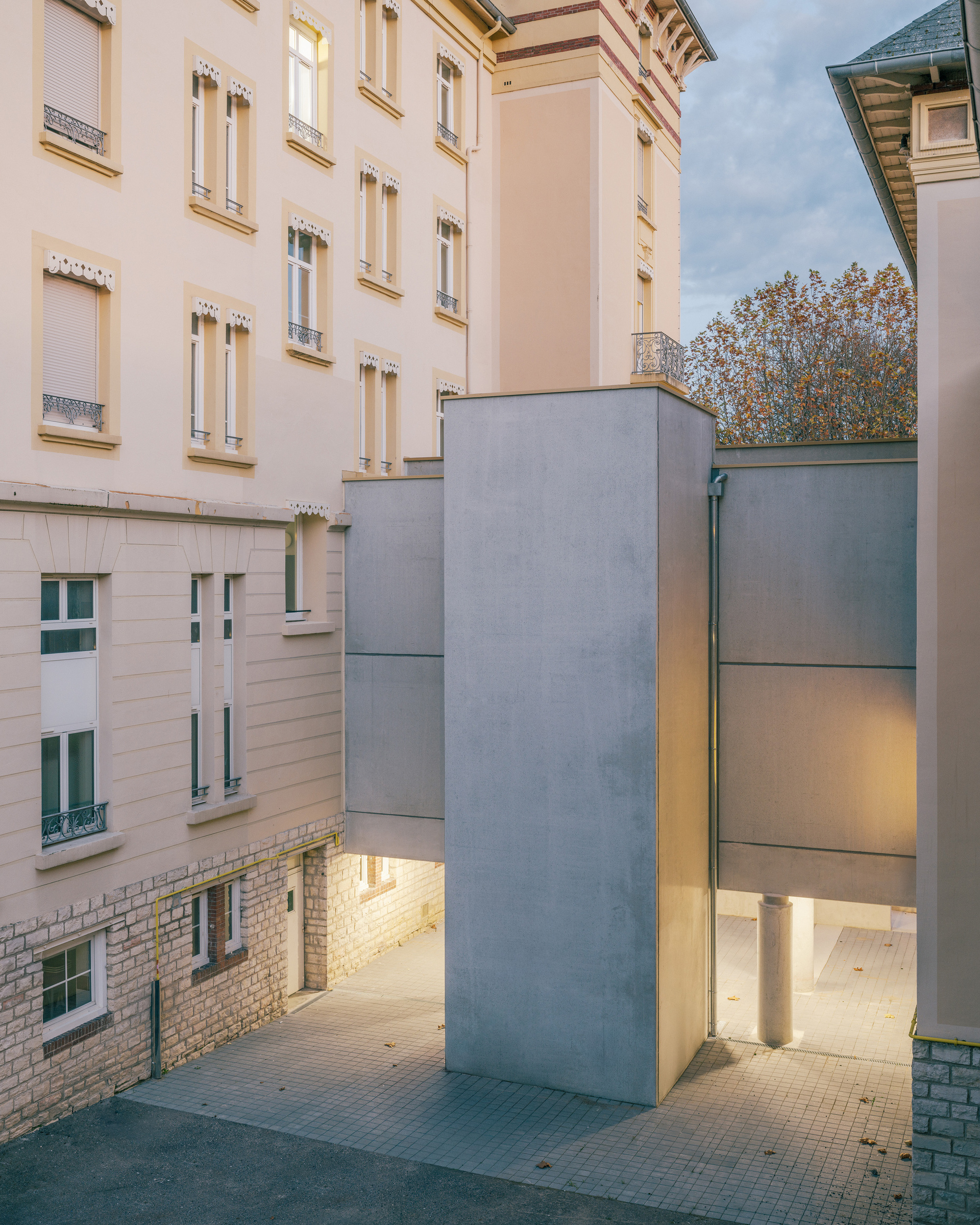 Courtyard view showing a grey fiber-cement volume inserted between existing residential buildings at dusk