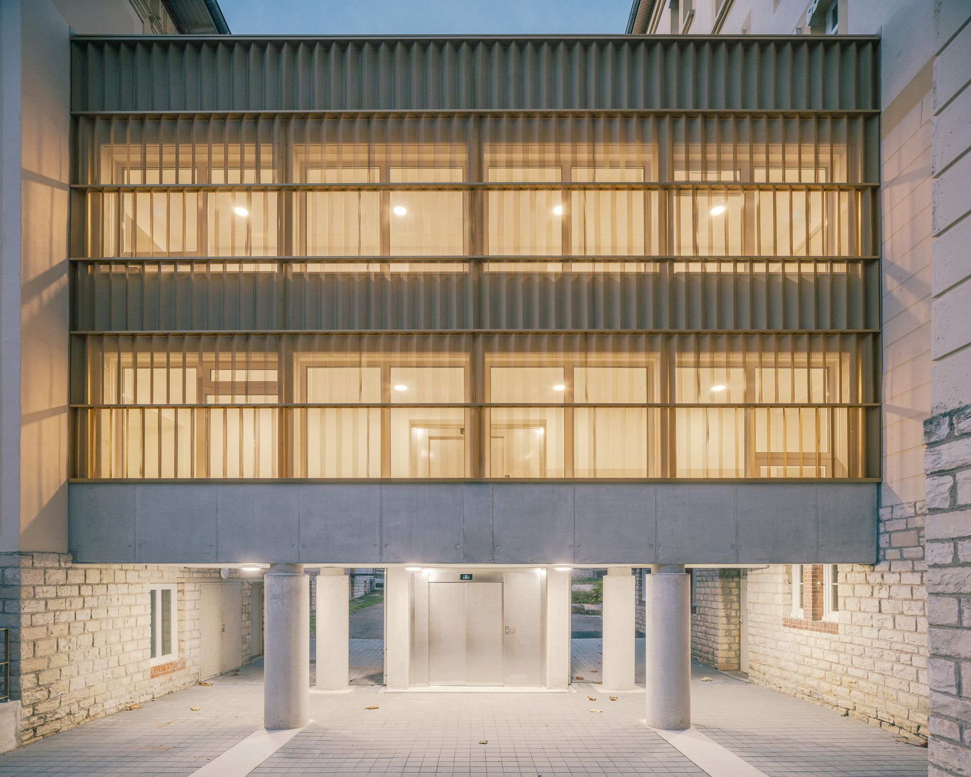 Illuminated timber-slatted facade with horizontal bands of glazing above a piloti entrance at twilight