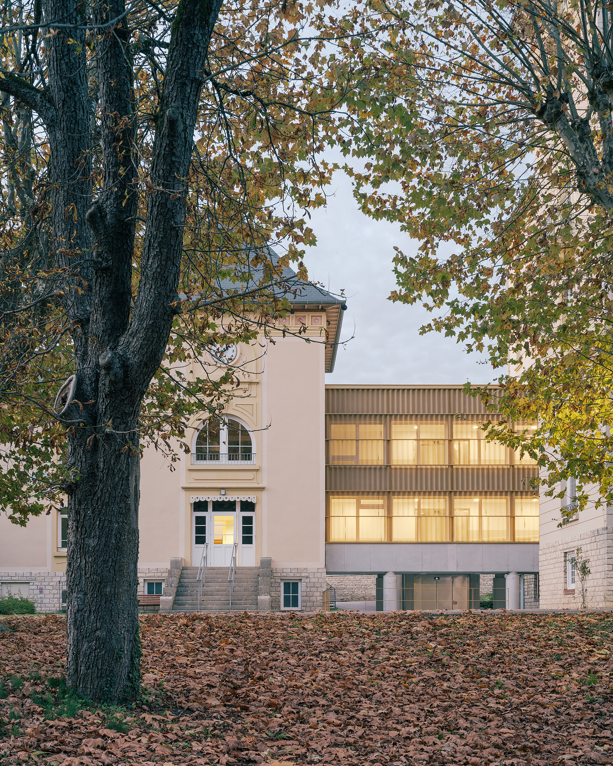 Illuminated corrugated metal facade addition between two pale plaster buildings framed by autumn foliage