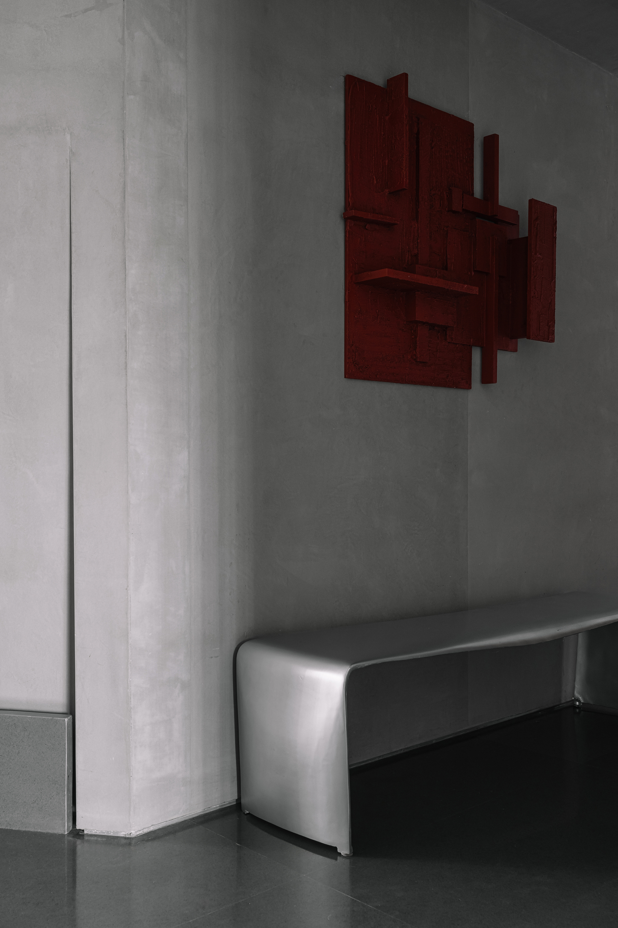 Detail of concrete wall corner with deep red sculptural wall piece above upholstered bench