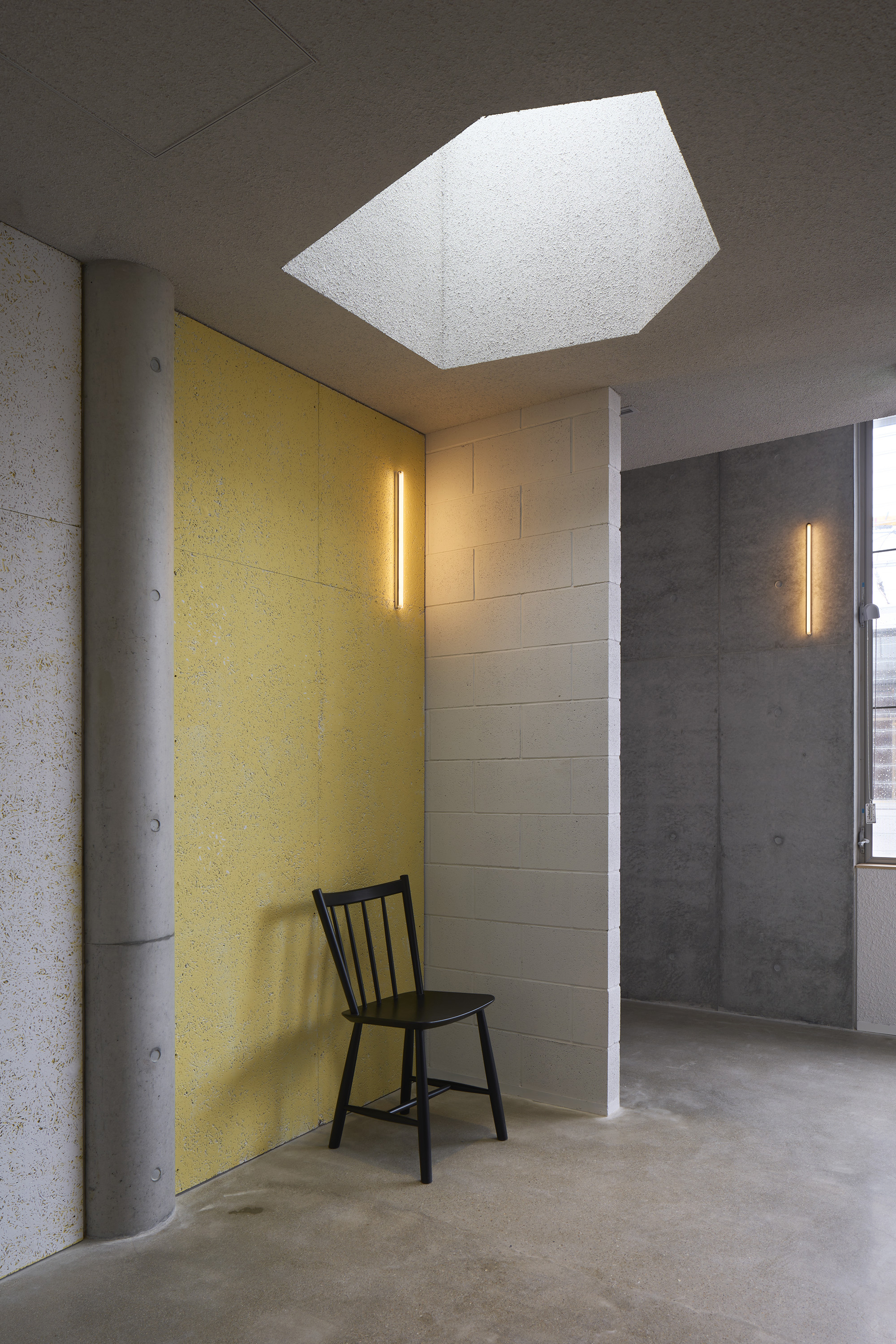 Pentagonal skylight opening in a dark grey ceiling above a yellow acoustic panel and black chair