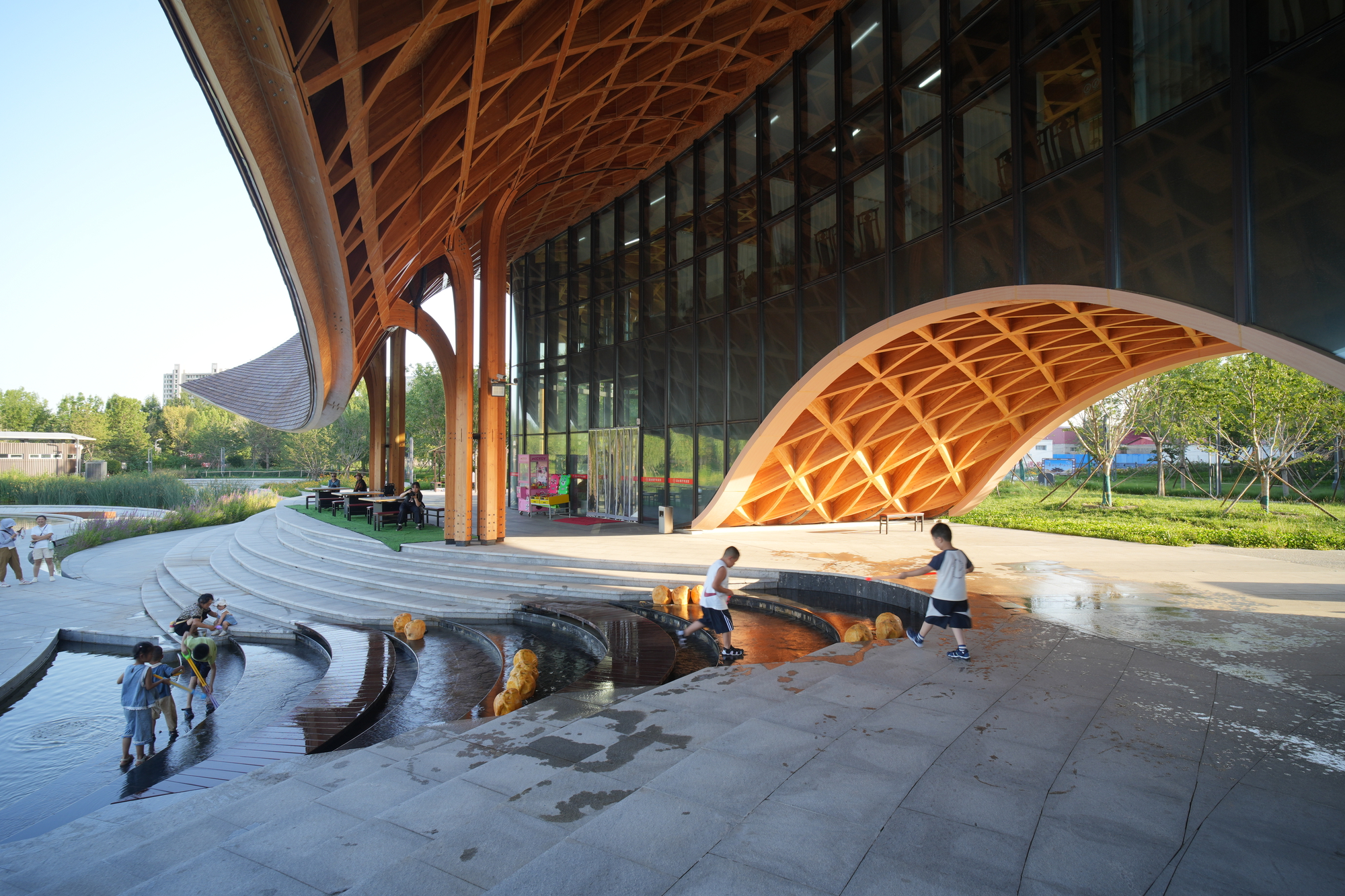 Children playing in the shallow water feature below the covered terrace with timber gridshell canopy overhead