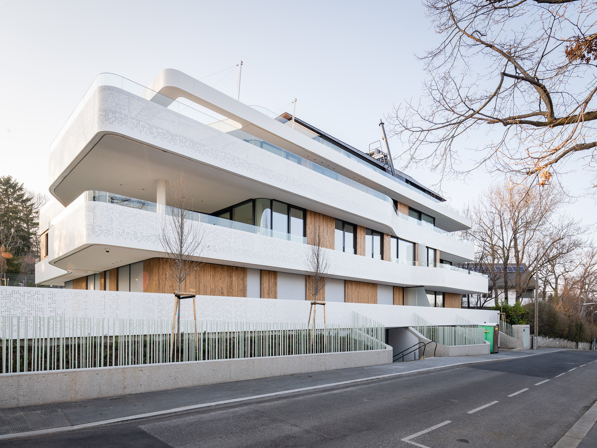 Side facade showing stacked terraces with perforated metal railings and concrete base wall from the roadway