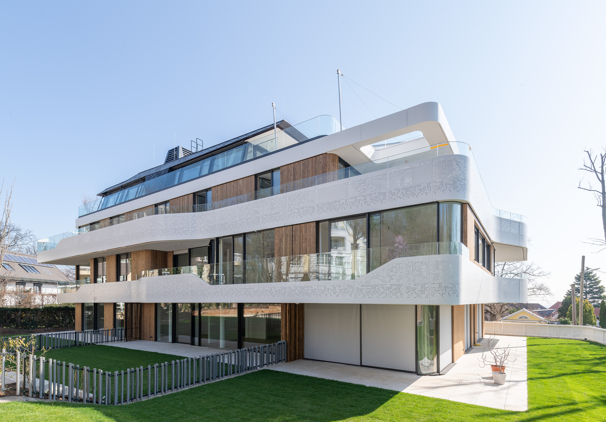 Corner view of the wraparound facade with perforated concrete balconies and timber panels above the lawn