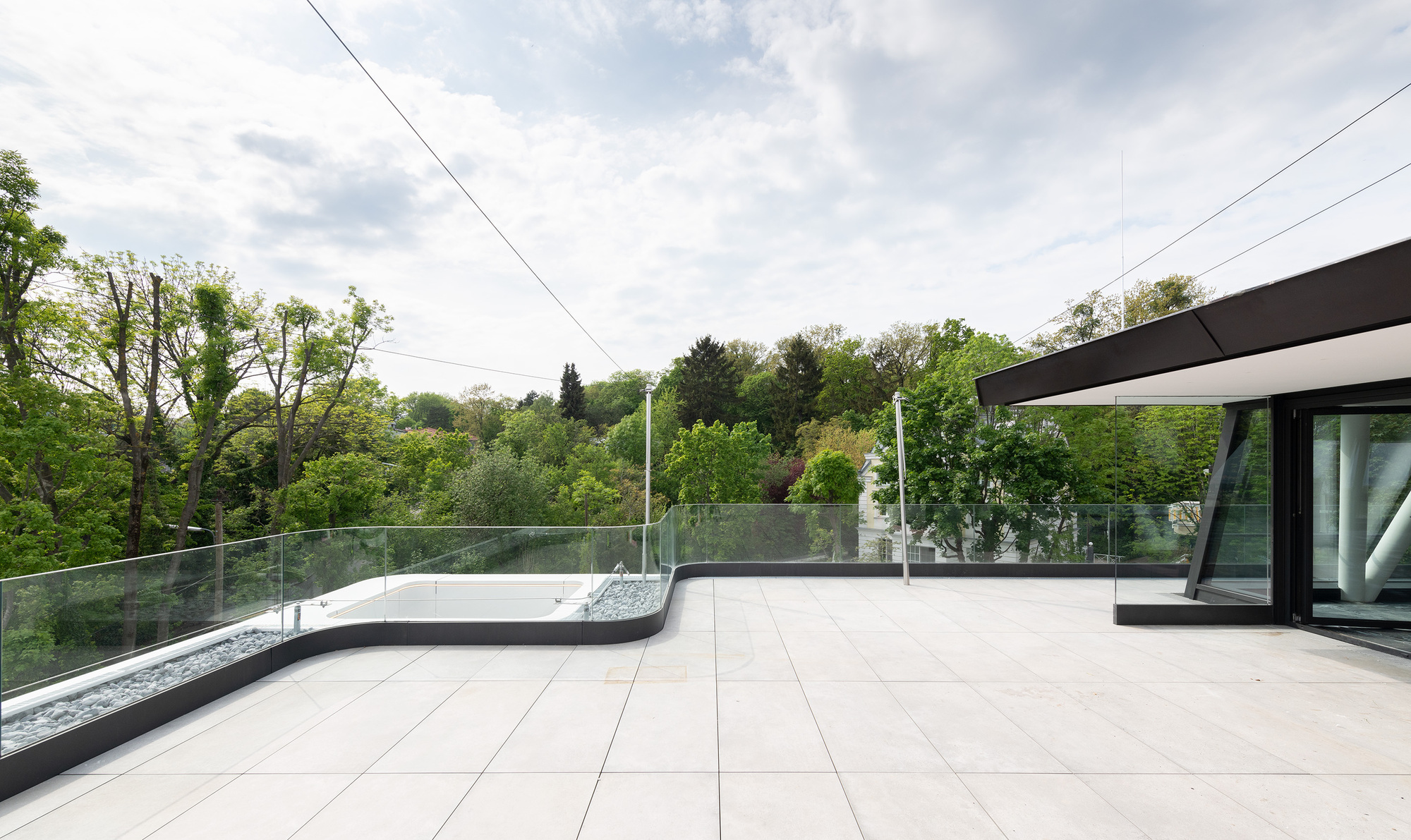 Rooftop terrace with pale pavers, glass balustrade, and tree canopy under cloudy sky