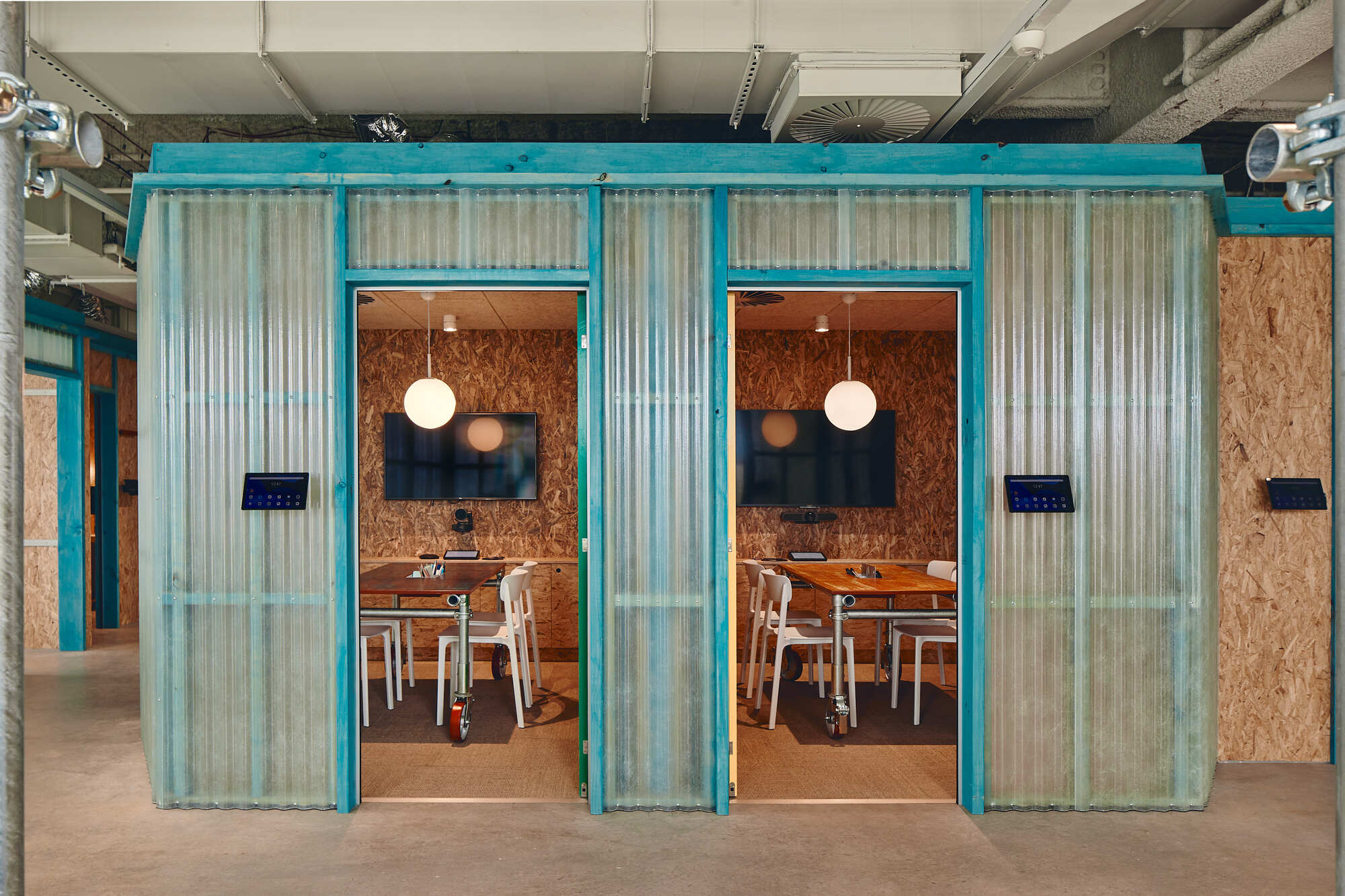 Meeting pods with turquoise corrugated metal frames and translucent curtains beneath exposed ceiling ducts