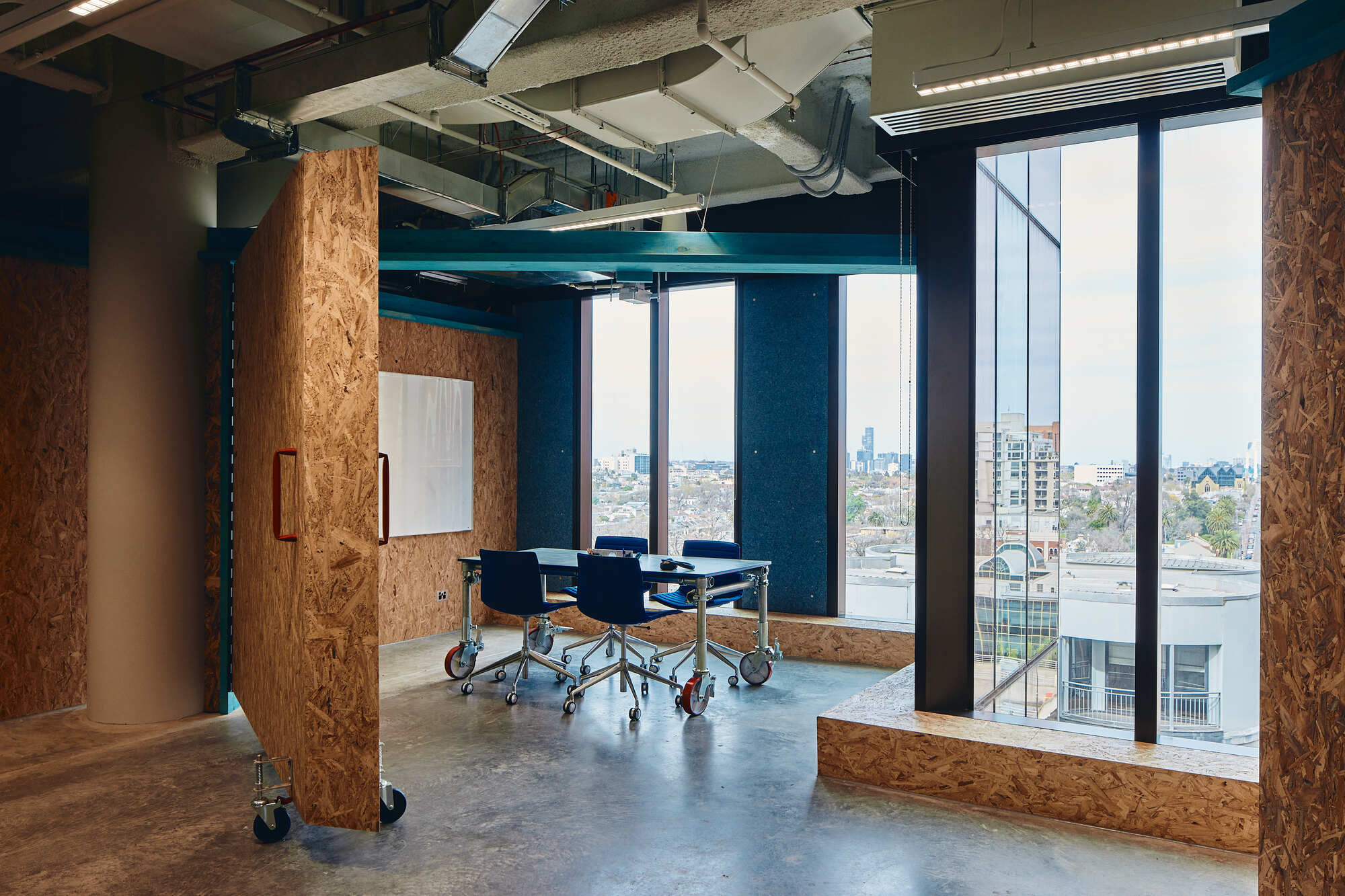 Meeting space enclosed by oriented strand board partitions on castors with floor-to-ceiling glazing beyond