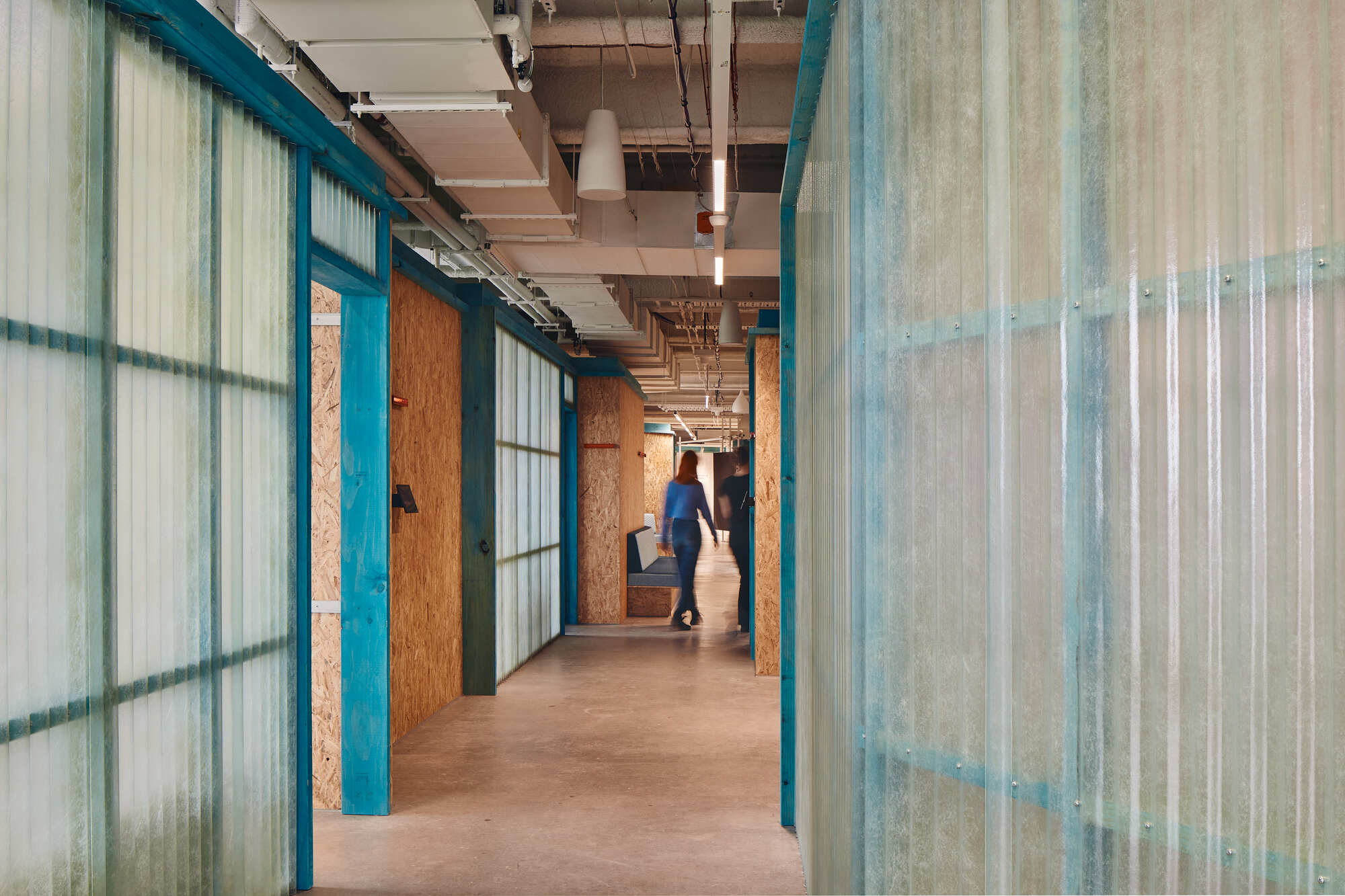 Corridor lined with translucent corrugated panels and oriented strand board partitions as a person walks through