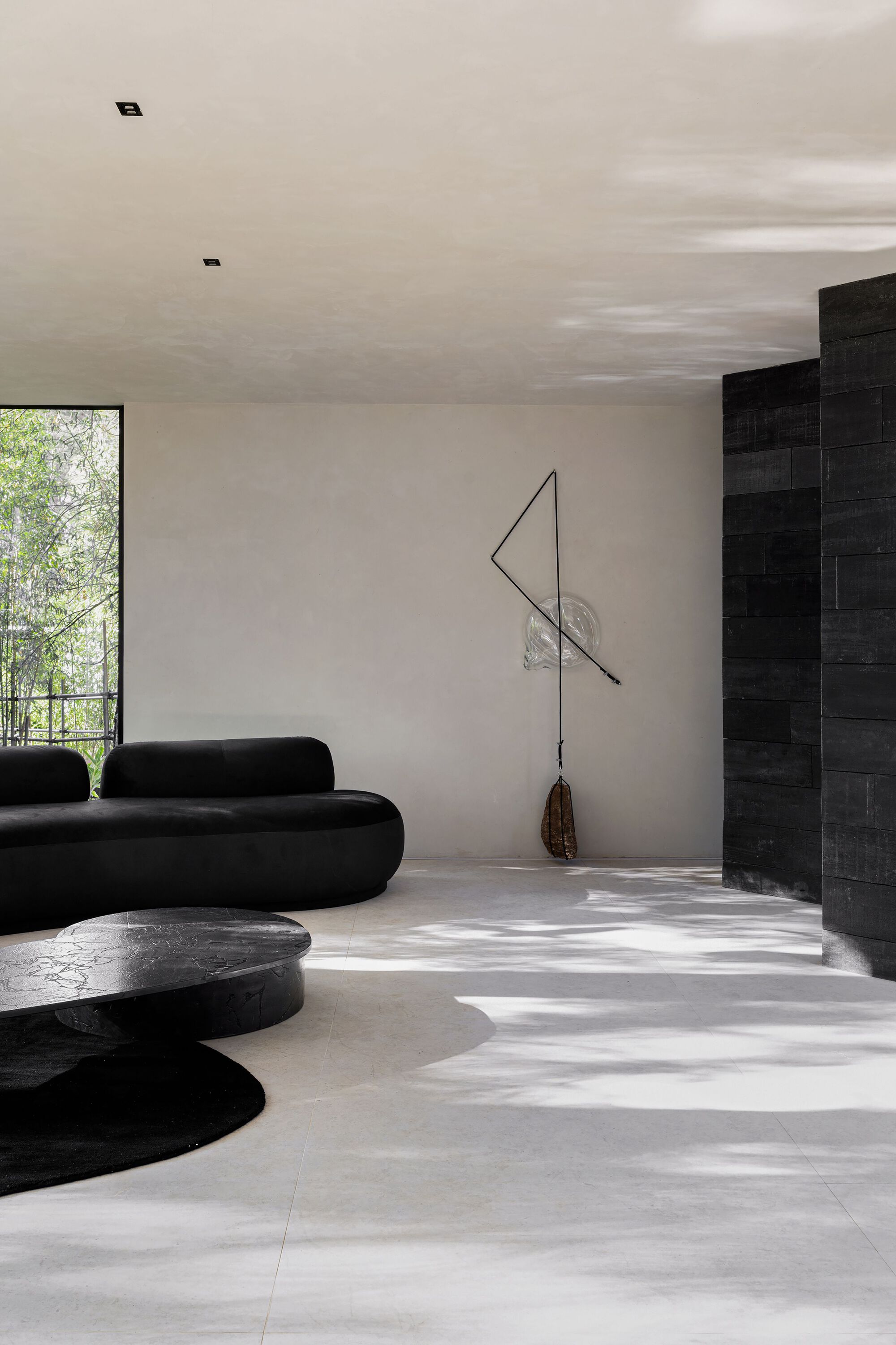 Living space with sculptural seating and dappled sunlight on concrete floor beside a black tiled wall