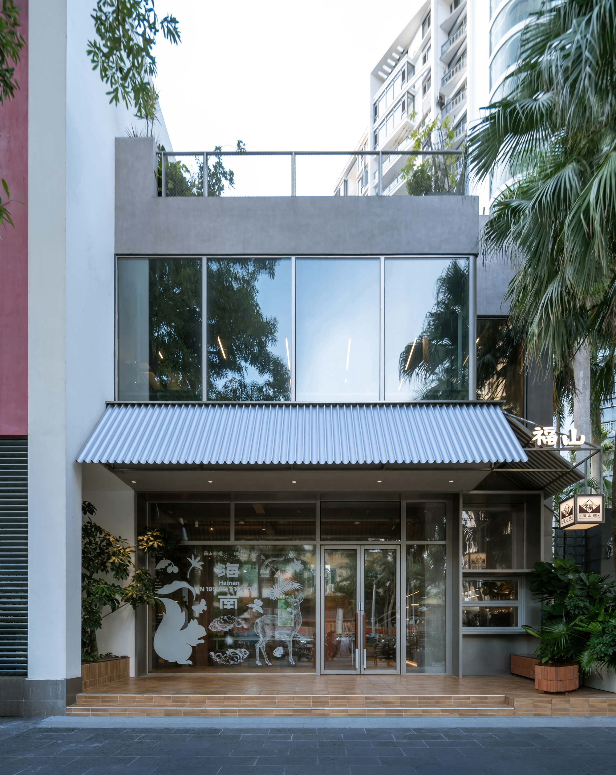 Street facade with corrugated metal canopy and ground-floor glazing framed by palms and residential towers