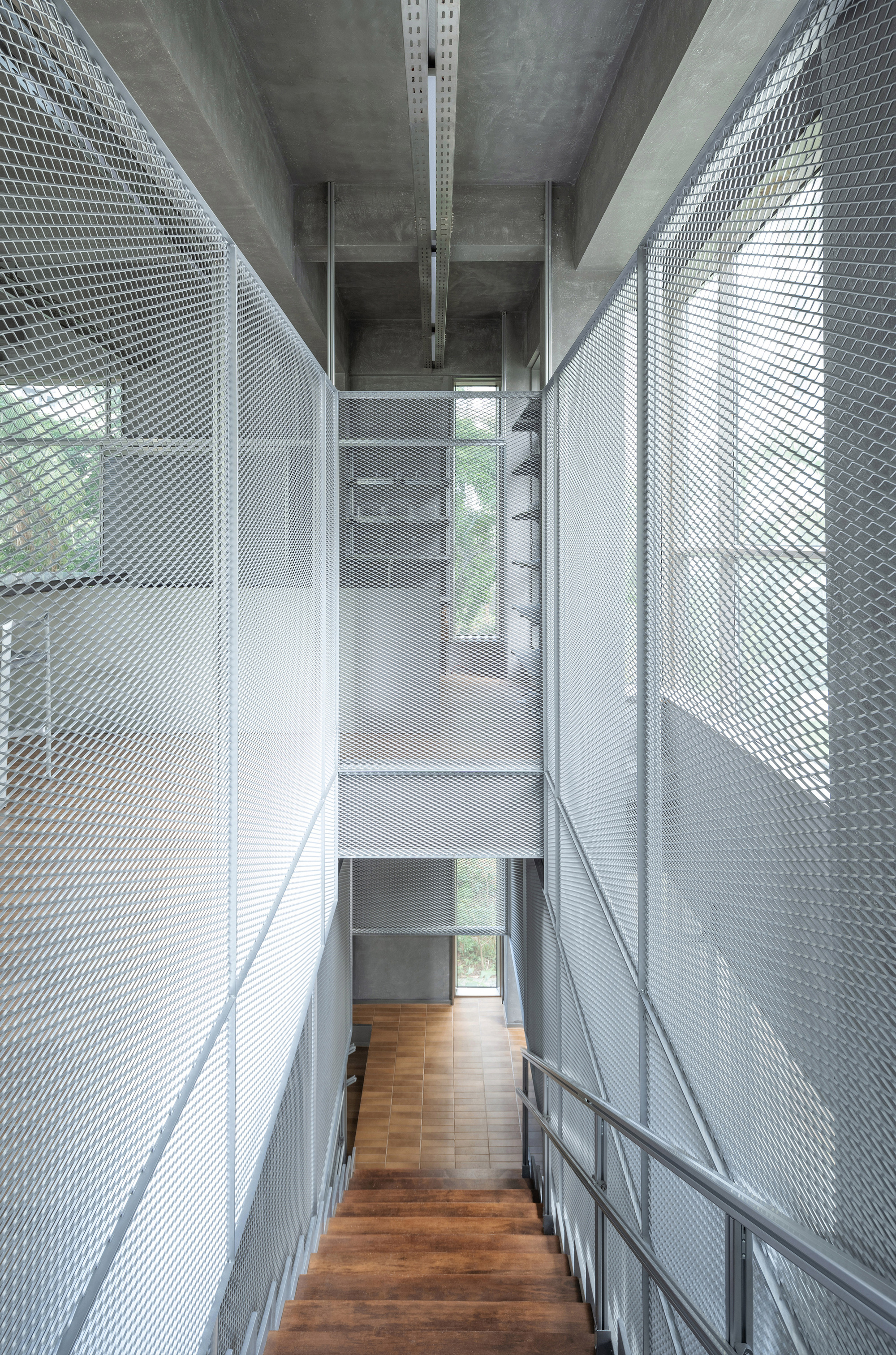 View down timber staircase flanked by expanded metal mesh screens filtering daylight through concrete beams