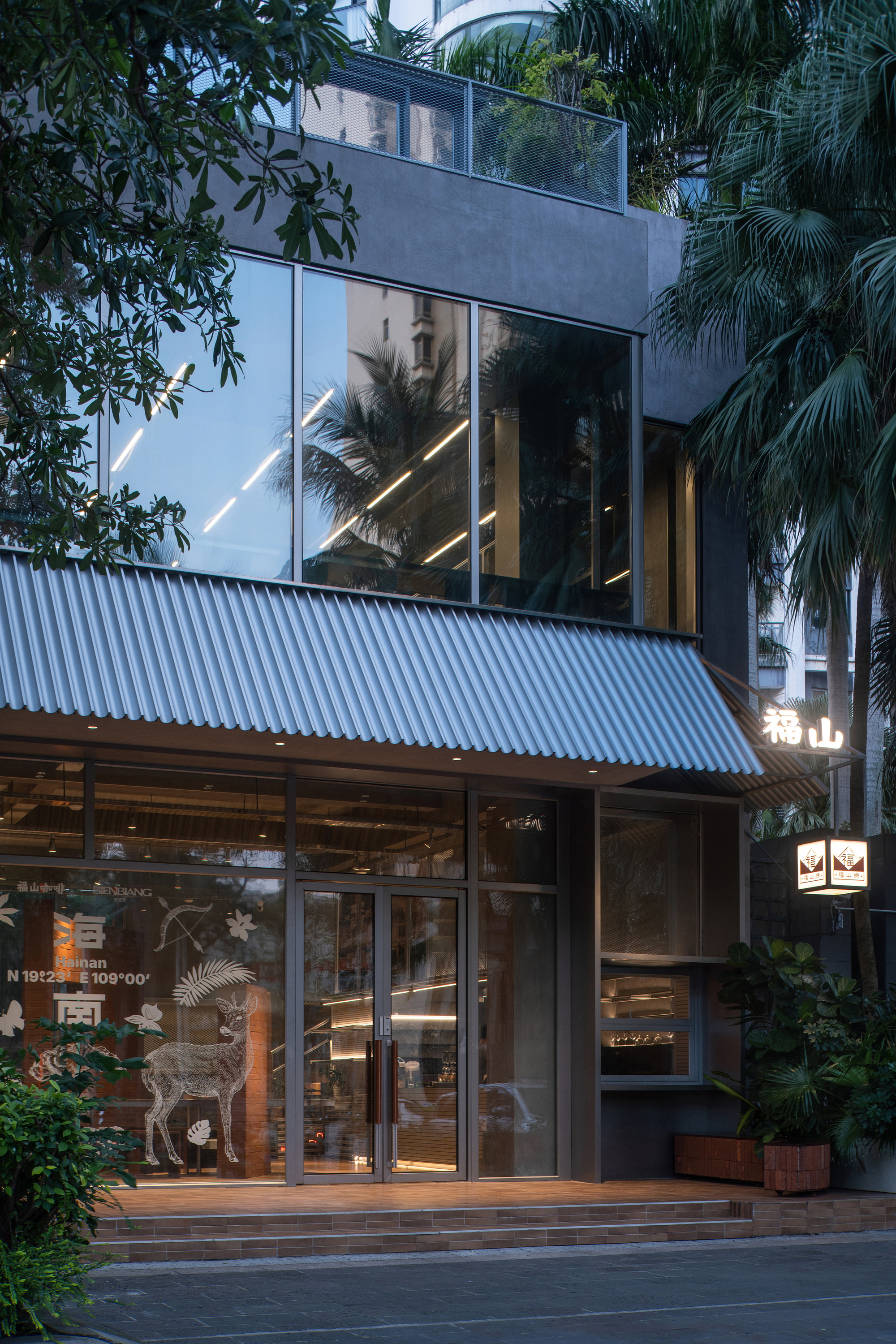 Three-story glazed facade with corrugated metal canopy framed by palm trees at dusk