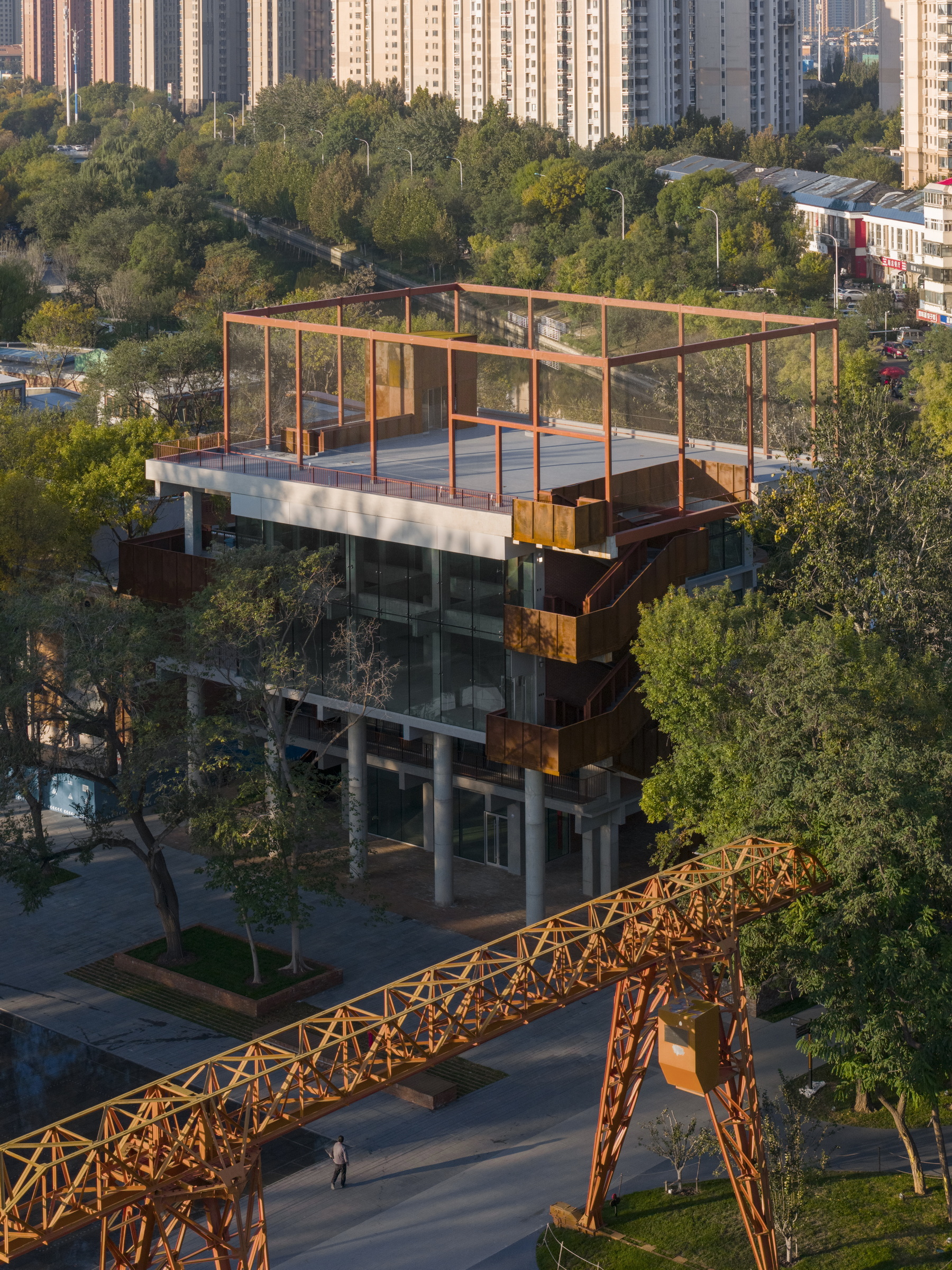 Aerial view of the rooftop terrace with red steel frame surrounded by park greenery and residential towers
