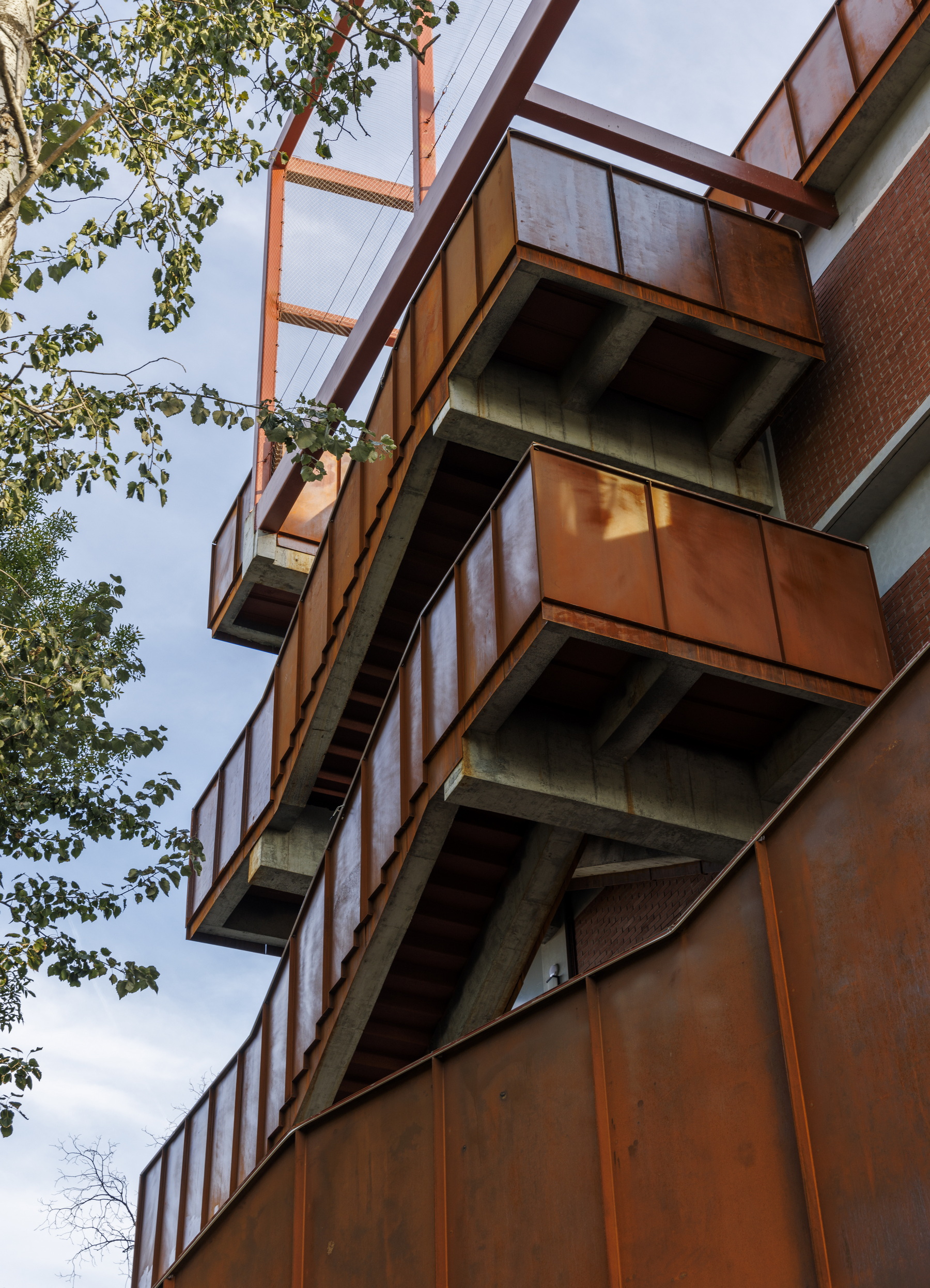 Cantilevered balconies clad in weathered steel with exposed concrete floor slabs and tree branches overhead