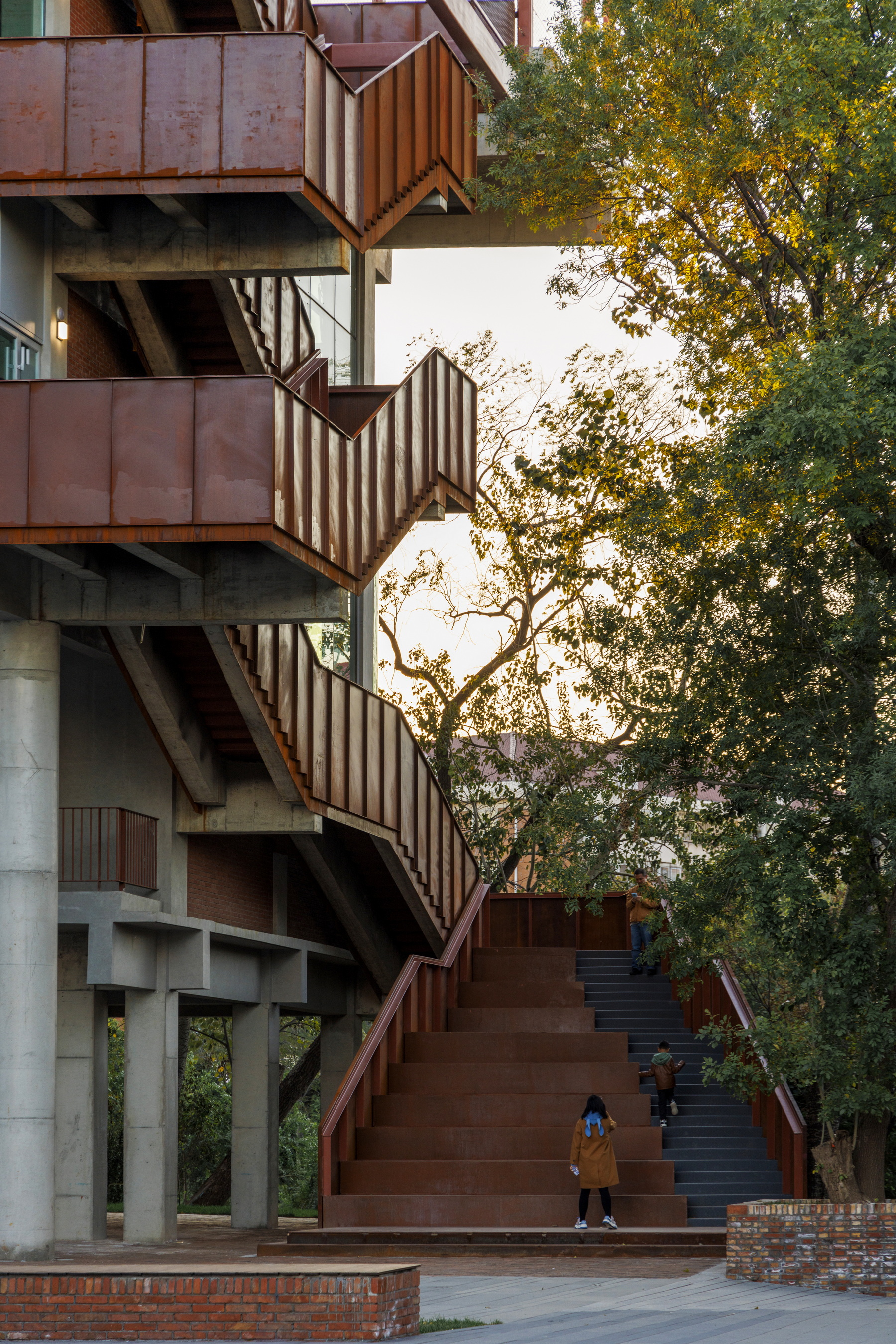 Wide exterior staircase with weathering steel cladding and concrete columns under trees in warm afternoon light