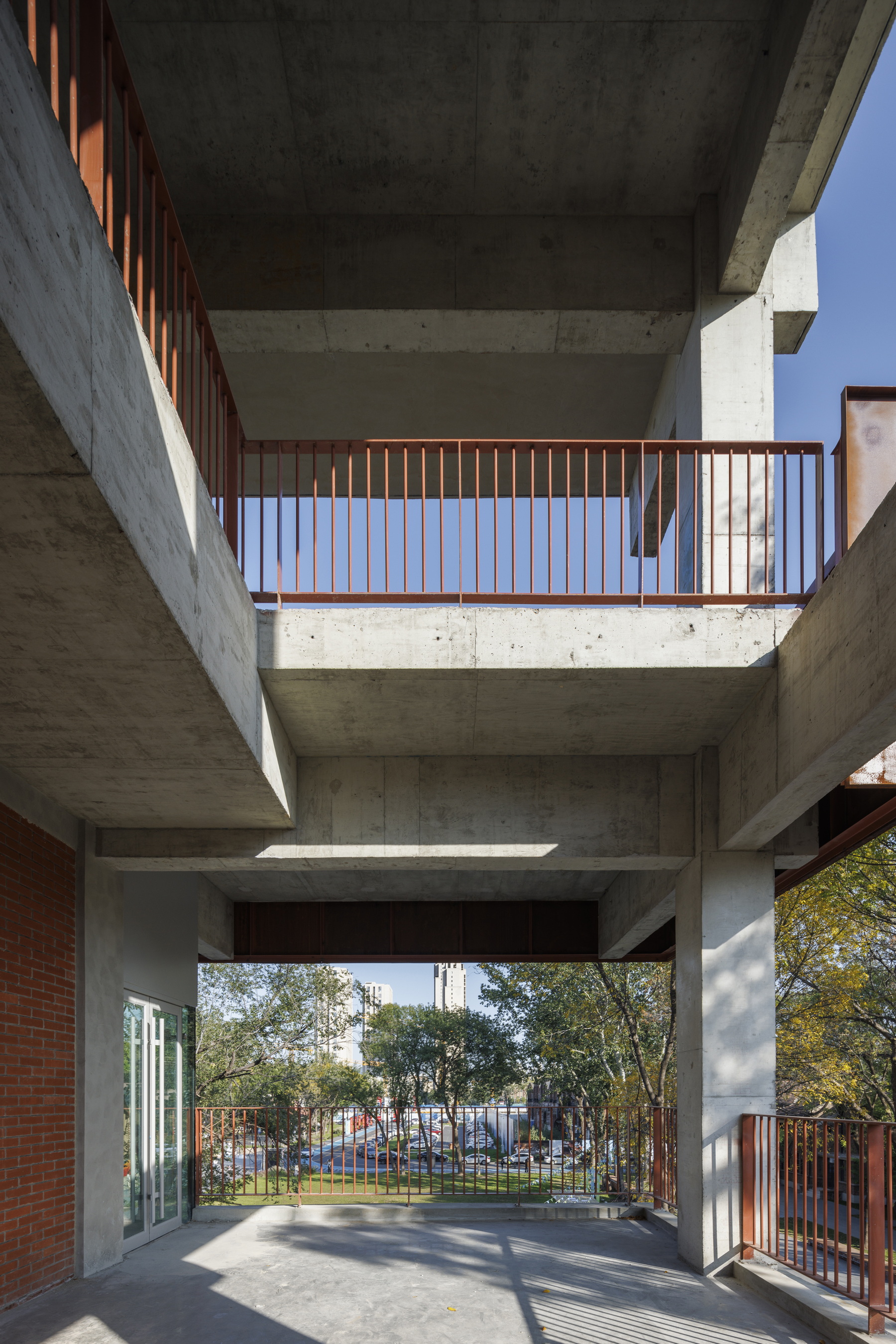 Ground level view through a concrete pilotis passage with steel balcony railings framing a planted courtyard beyond