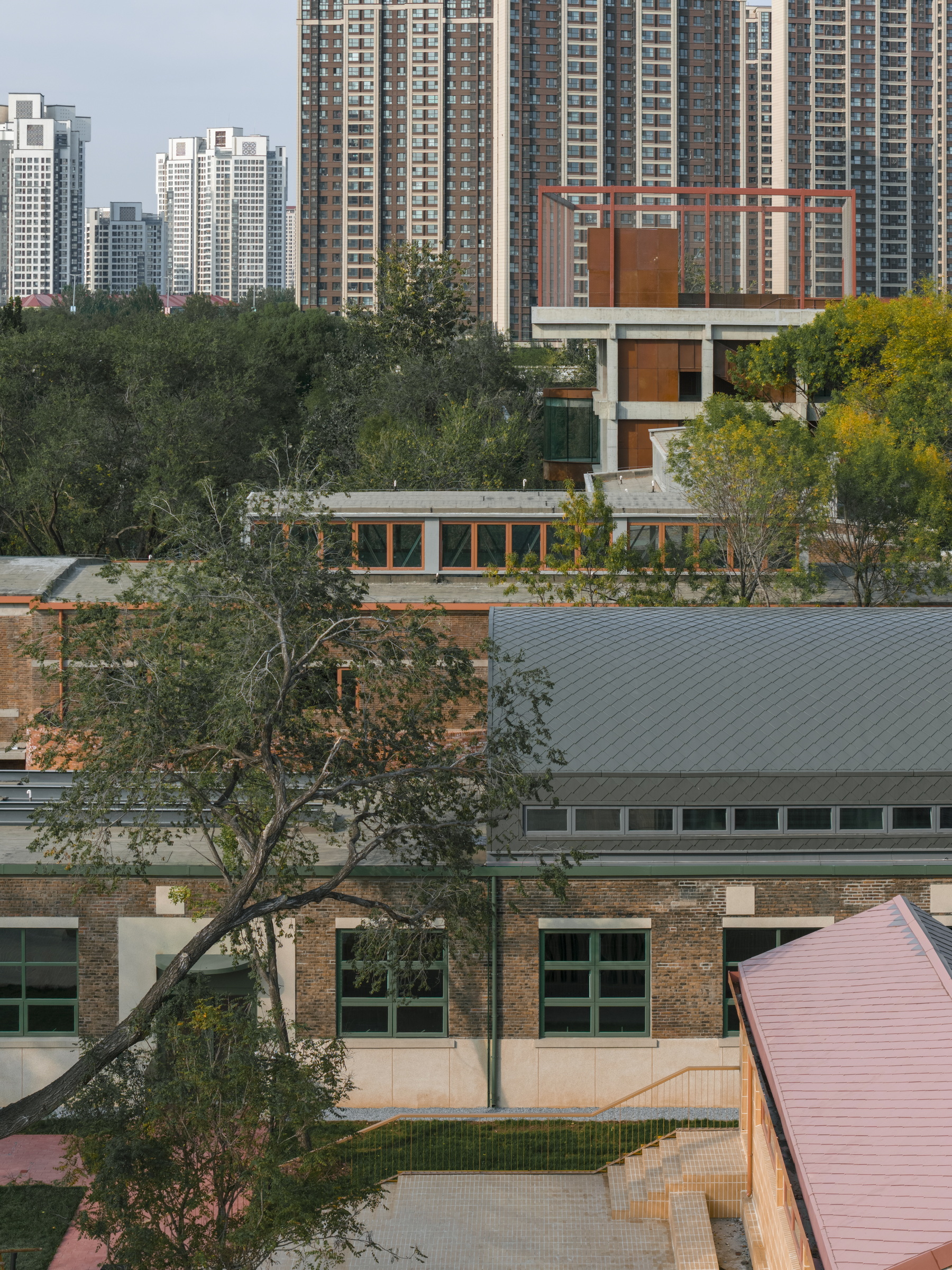 Elevated tower with rust-colored metal screens and glass walls emerging from treetops with apartment blocks behind