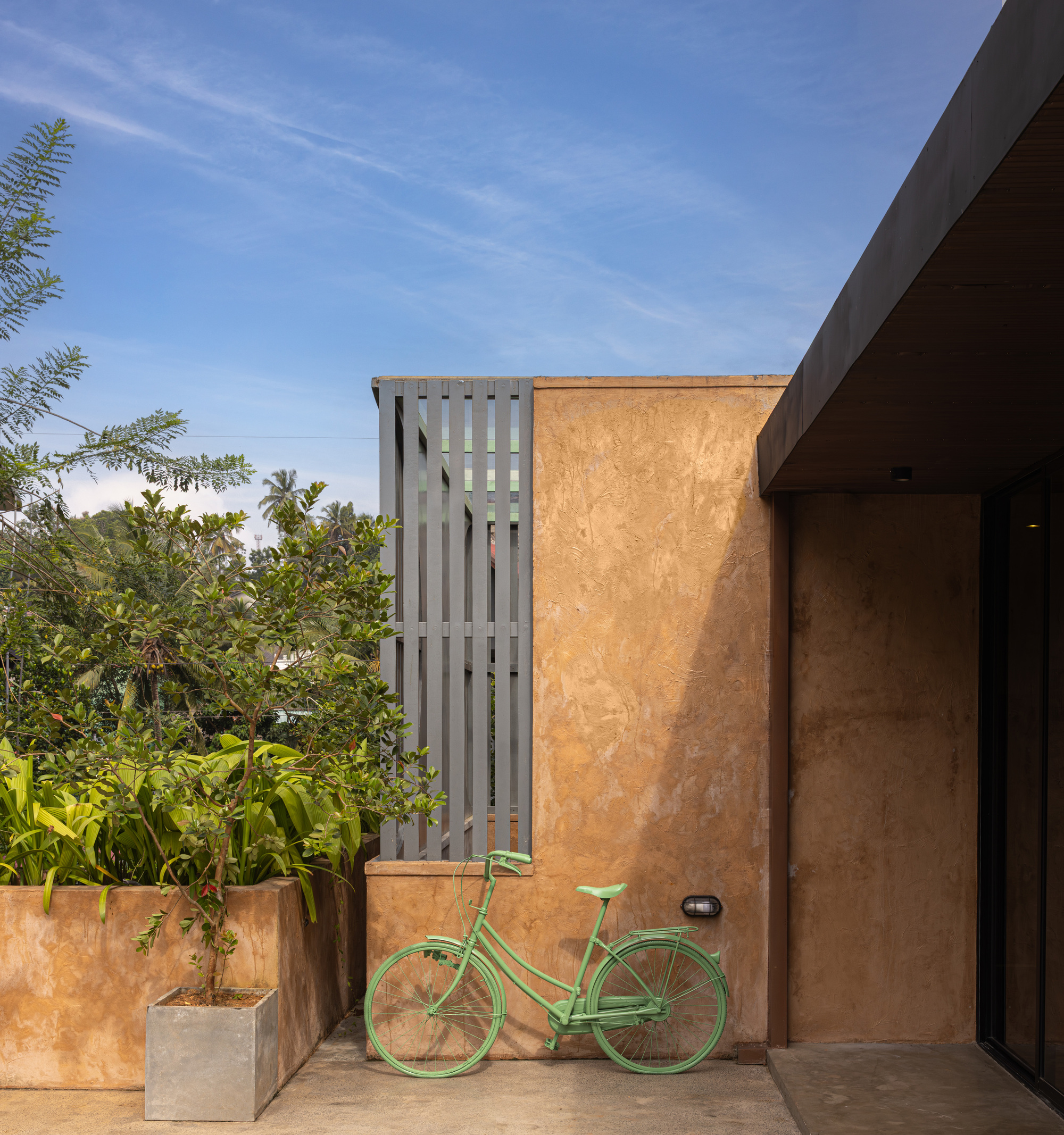 Courtyard view with weathered ochre walls, vertical slatted screen, planted beds and a green bicycle under blue sky