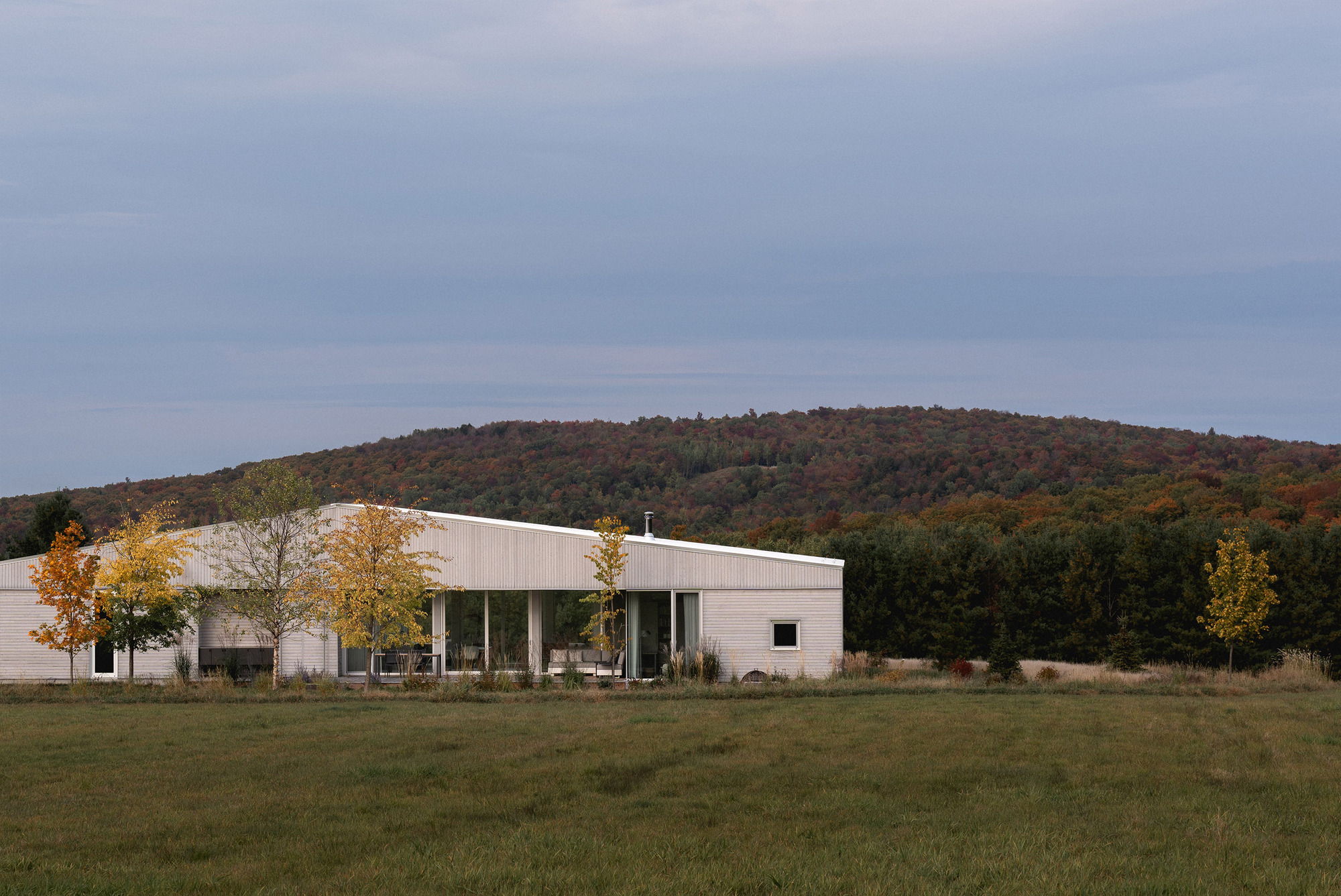 Long view of the white metal-clad residence set in an open meadow with forested hills beyond