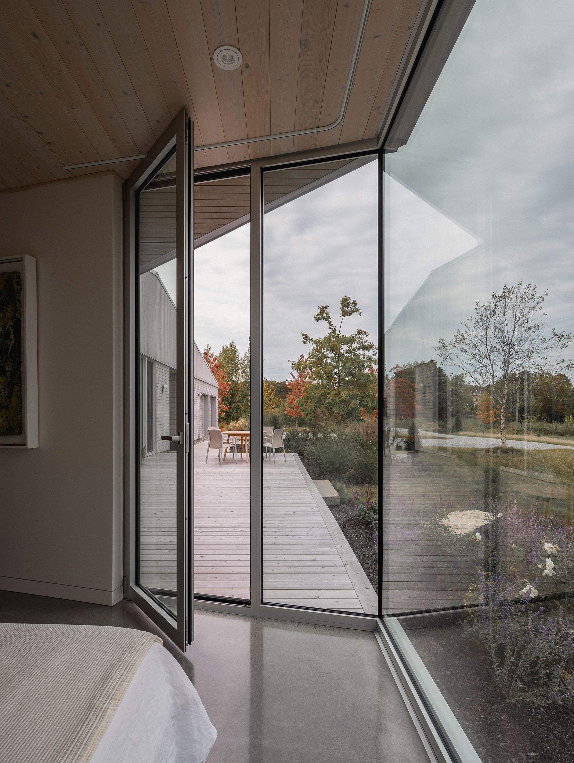 Pivoting glass door opens from bedroom onto timber deck framed by autumn foliage and cloudy sky