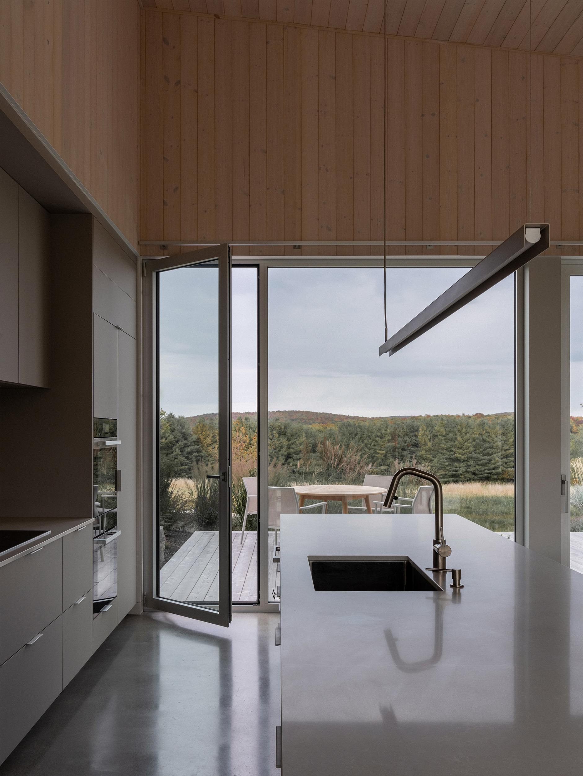 Kitchen island with integral sink facing narrow glass door and distant wooded hillside under overcast sky