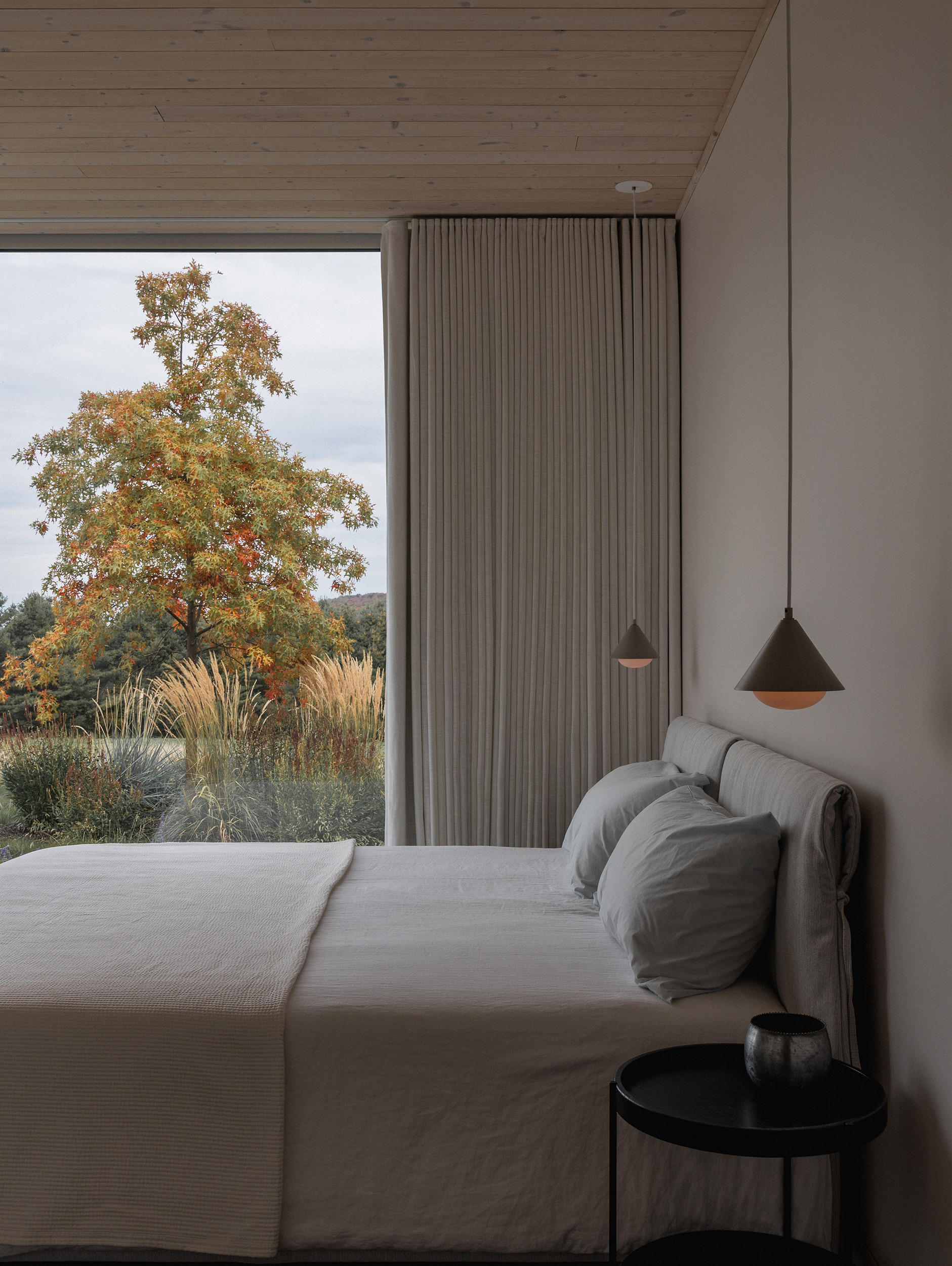 Bedroom corner with pendant lights and pleated curtain beside glazing overlooking grasses and an oak tree