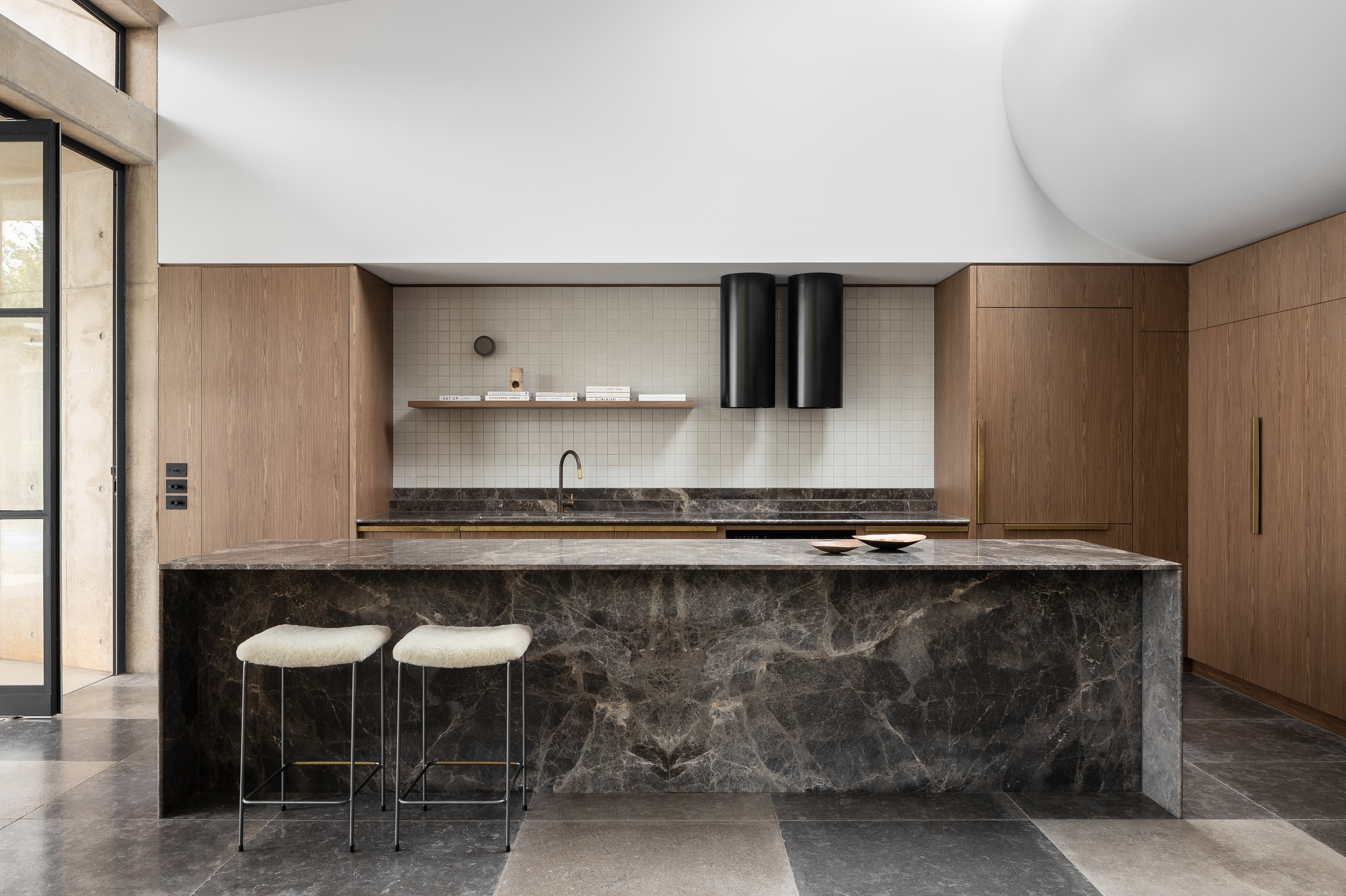 Kitchen island clad in dark stone with timber cabinetry and cylindrical black pendant lights above