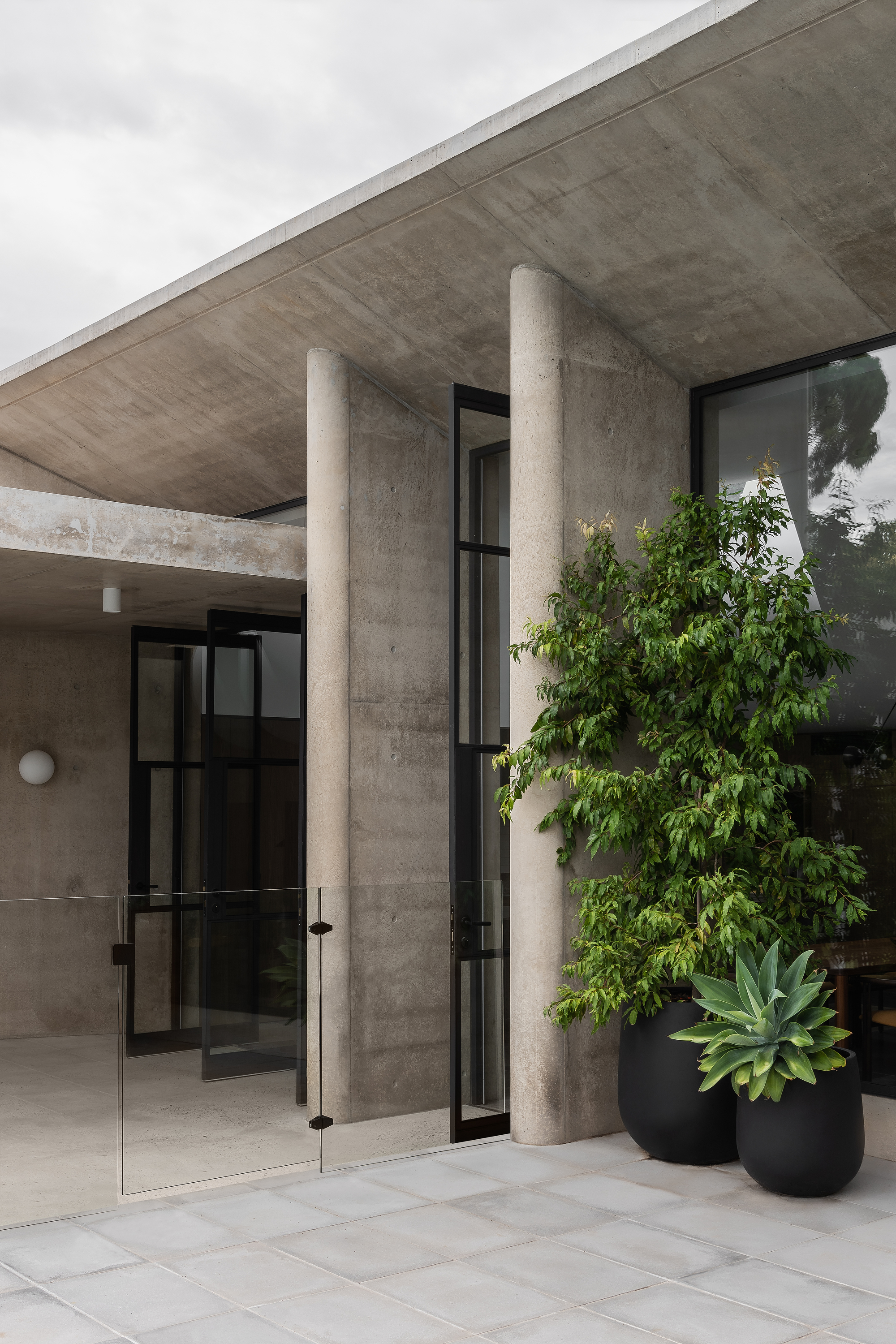 Covered entry porch with circular concrete columns and potted plants beneath an overcast sky