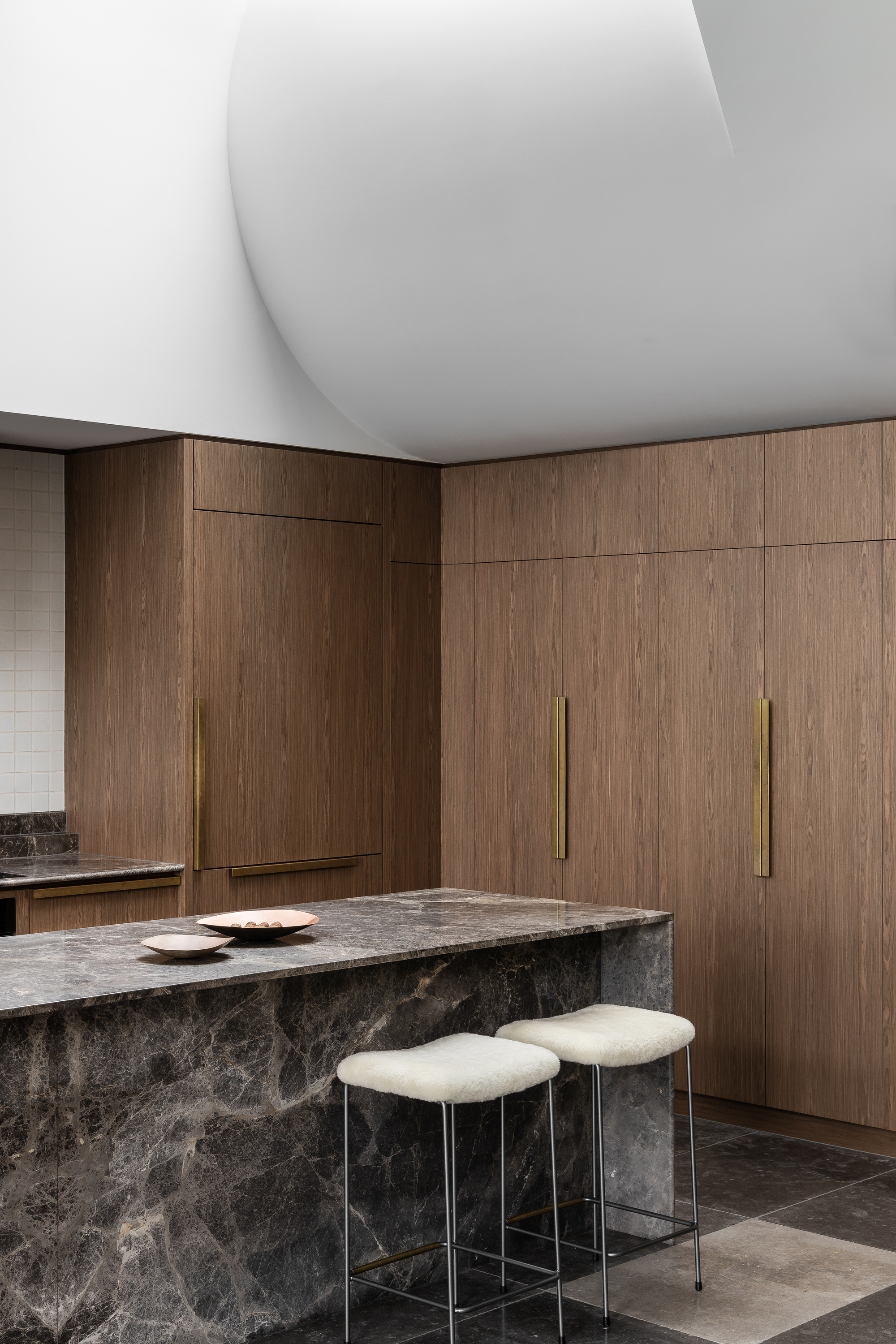 Dark stone island with brass-handled timber cabinetry and upholstered stools in natural light