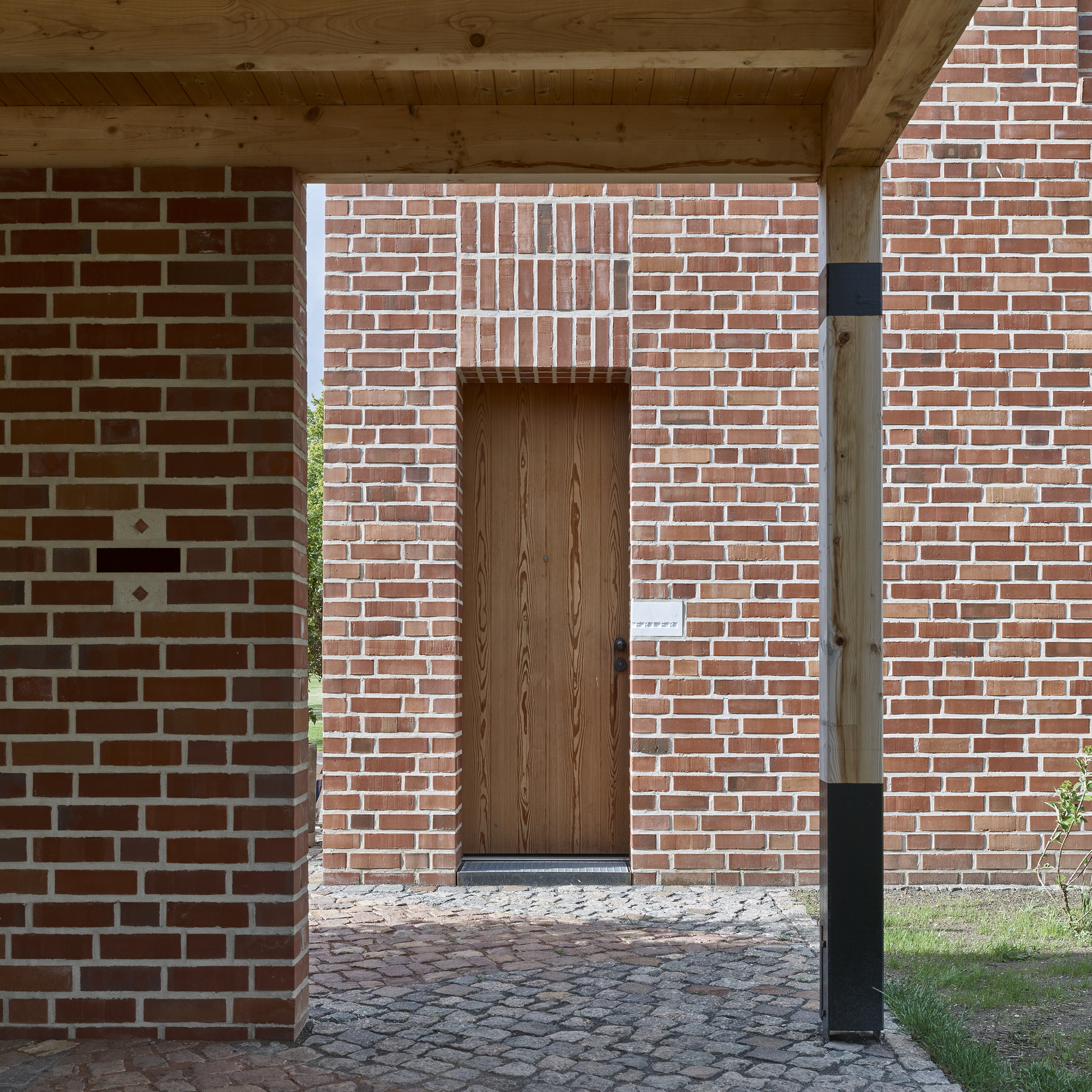 Timber entry door recessed in brick wall with exposed wood rafters overhead and cobblestone paving