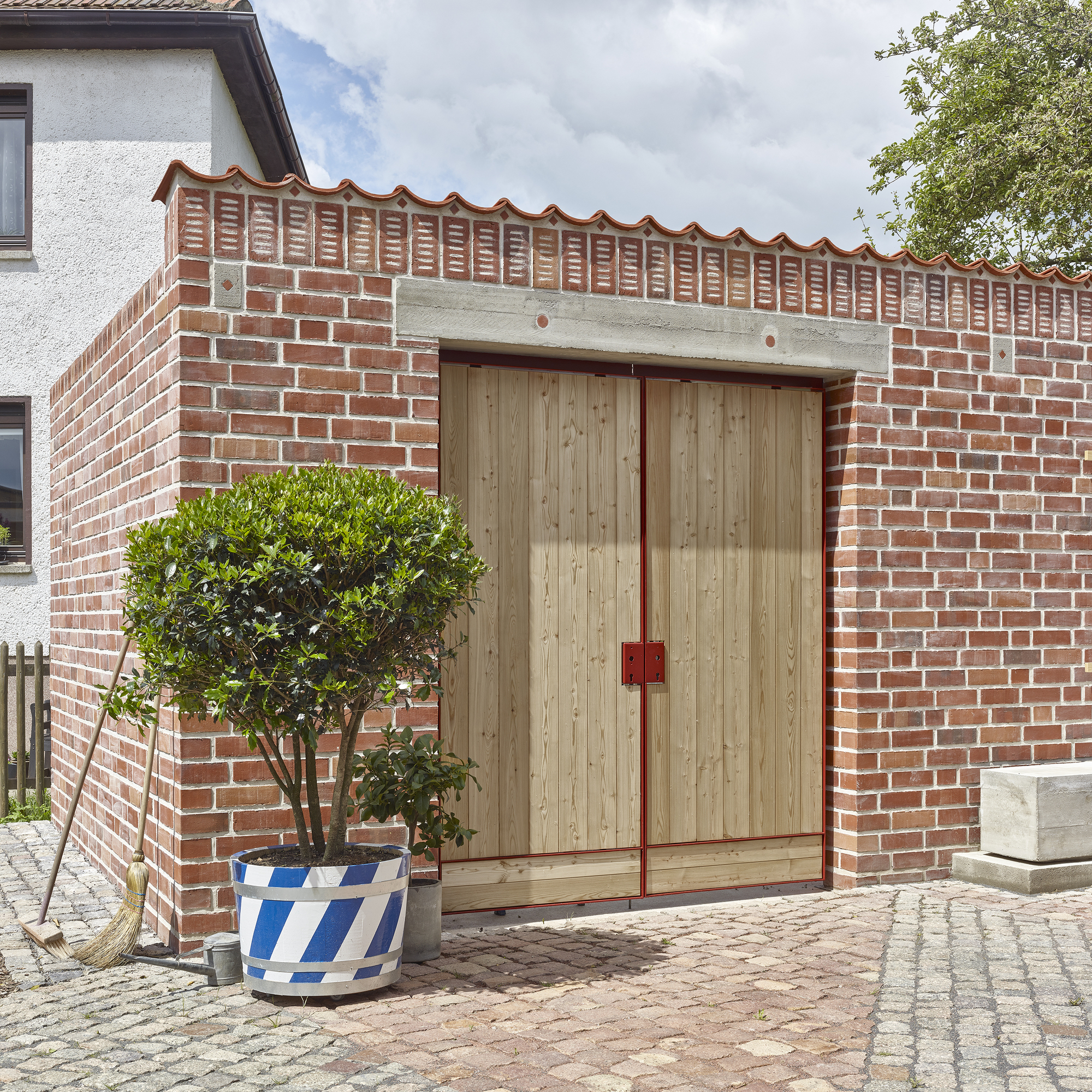 Street facade with double timber doors set below wavy terracotta coping and potted tree in foreground