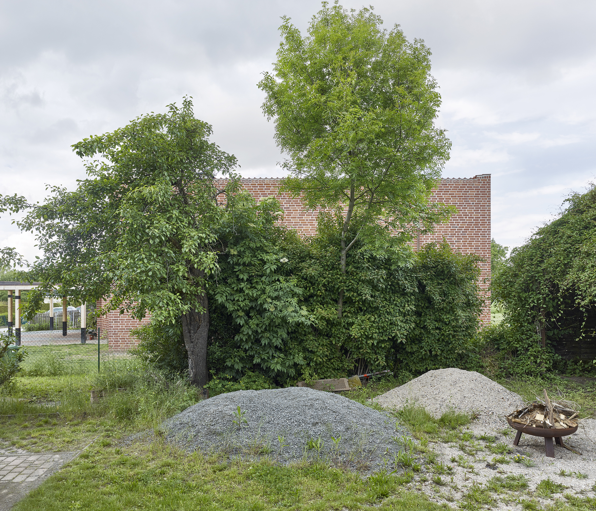 Distant view of the brick volume behind mature deciduous trees and gravel mounds under overcast skies