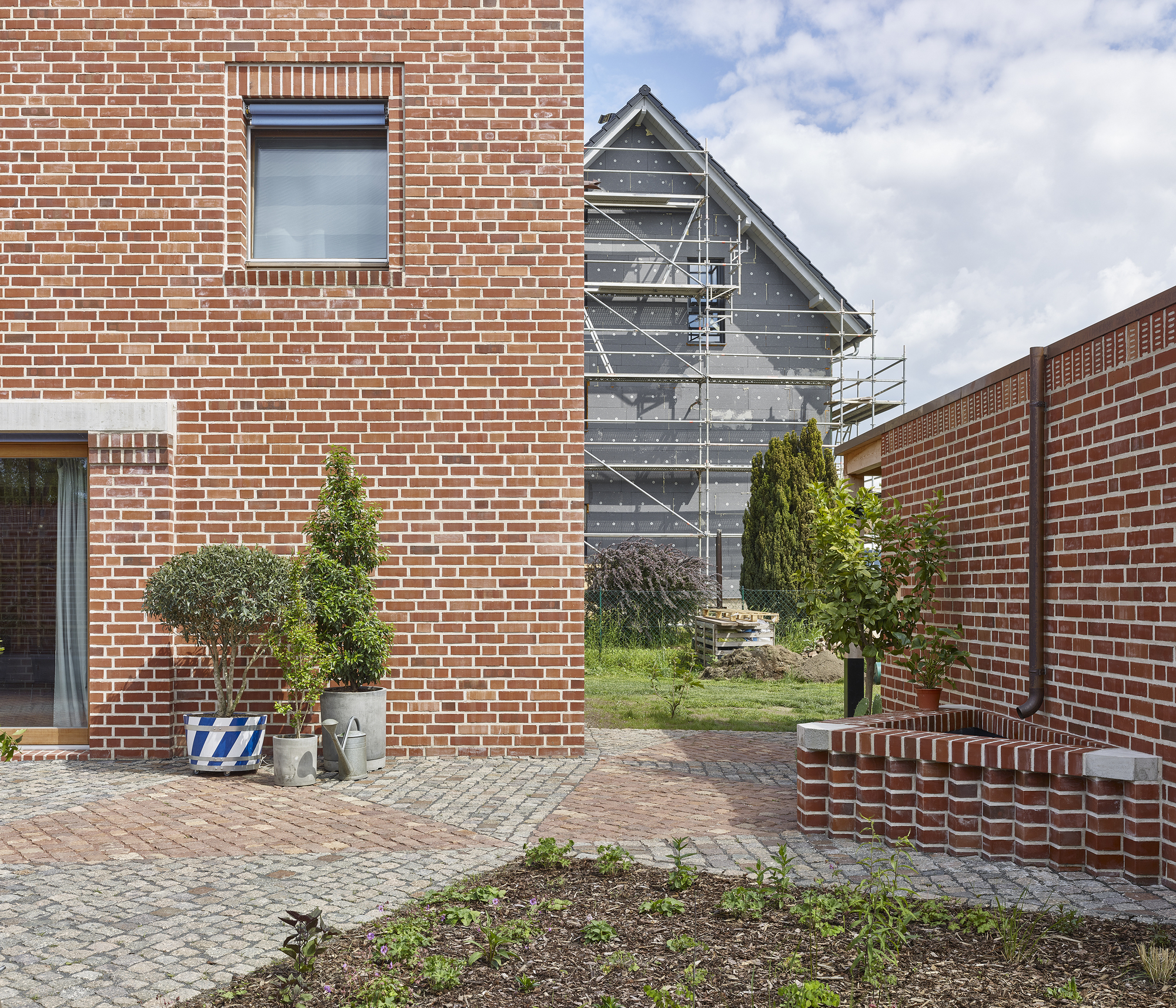 Brick-paved courtyard between red masonry walls with a dark metal gable structure visible beyond under cloudy sky