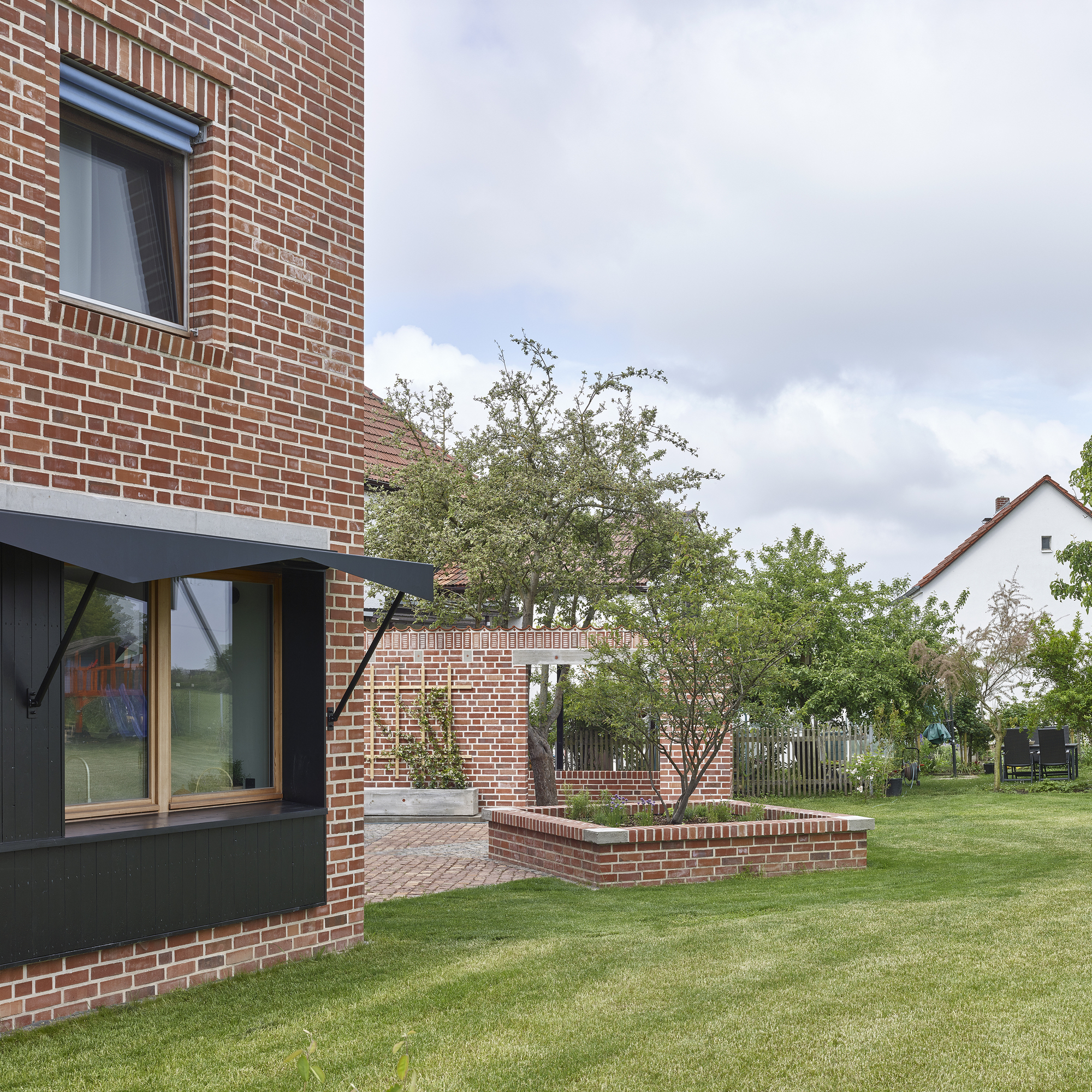 Red brick facade with cantilevered metal awning and planted brick planter beside a lawn with trees