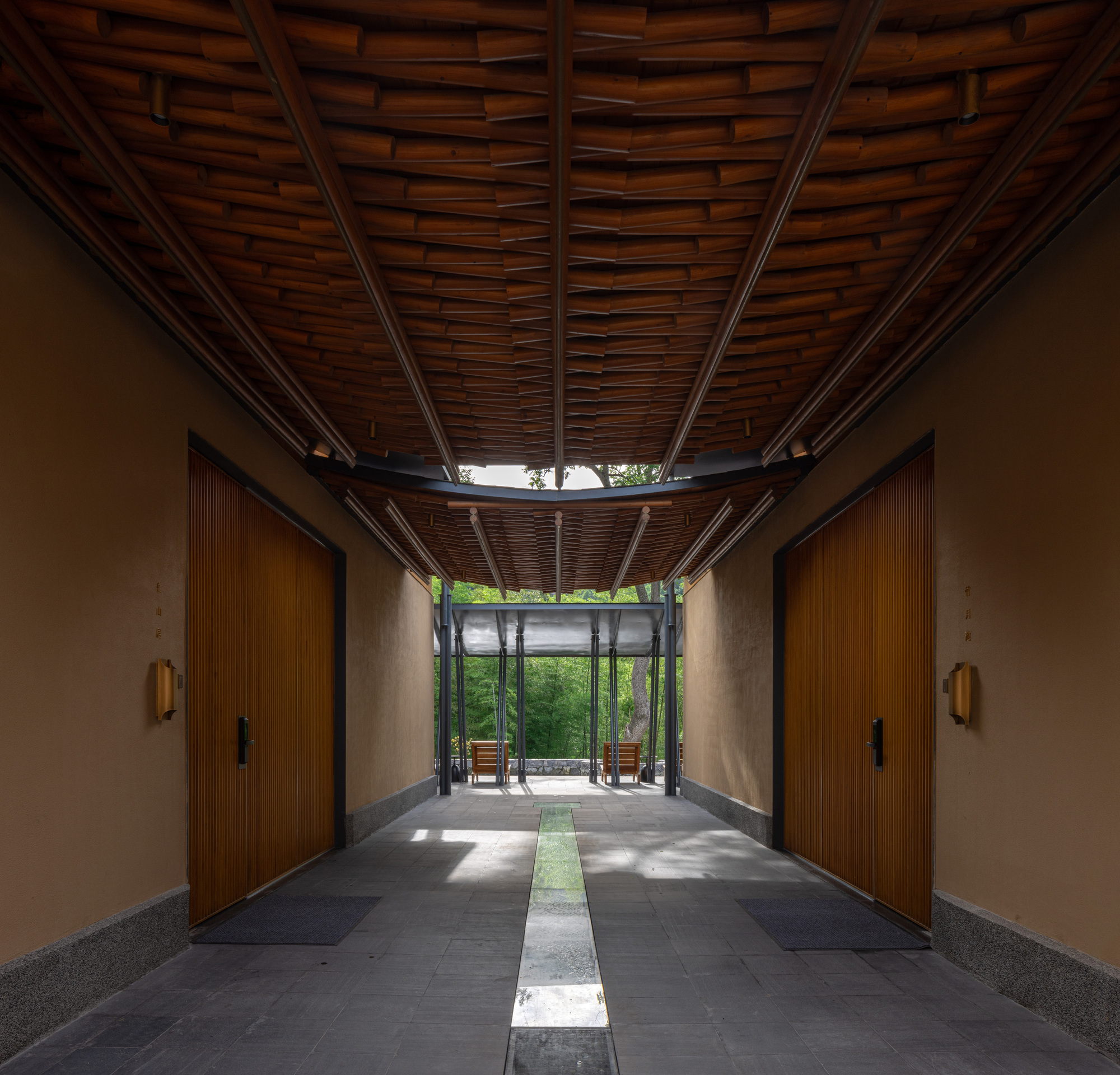 Entry corridor with woven timber ceiling, stone floor with water channel, and green trees visible beyond