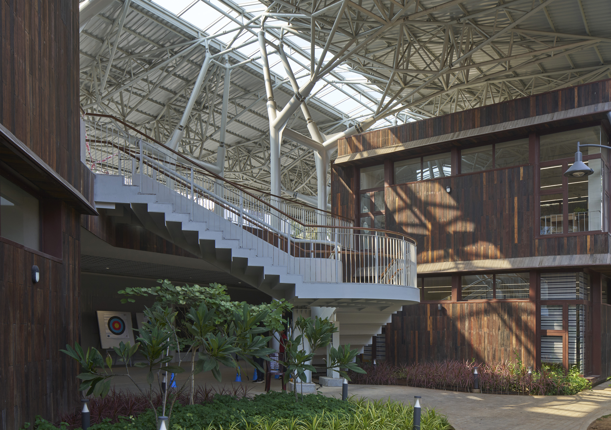Planted courtyard with a curving staircase connecting levels under dappled sunlight through the space frame