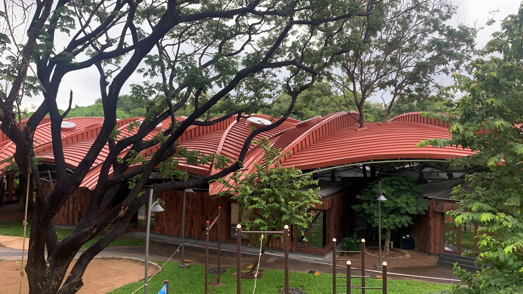 Undulating red metal roof canopy sheltering timber pavilions amid mature trees on a grassy lawn