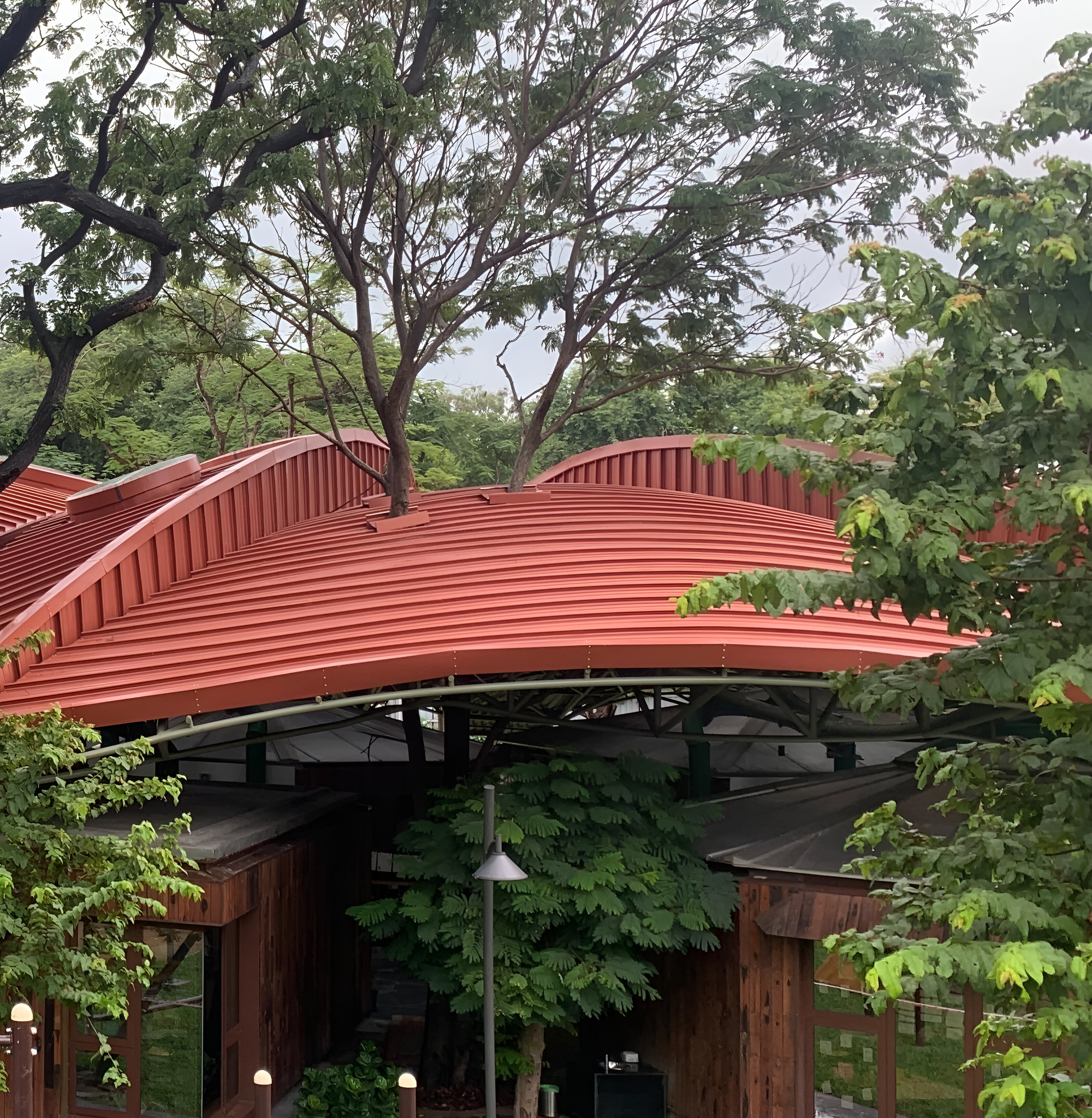 Section of the arching red metal roof with vertical ribbing sheltering an open pavilion surrounded by foliage