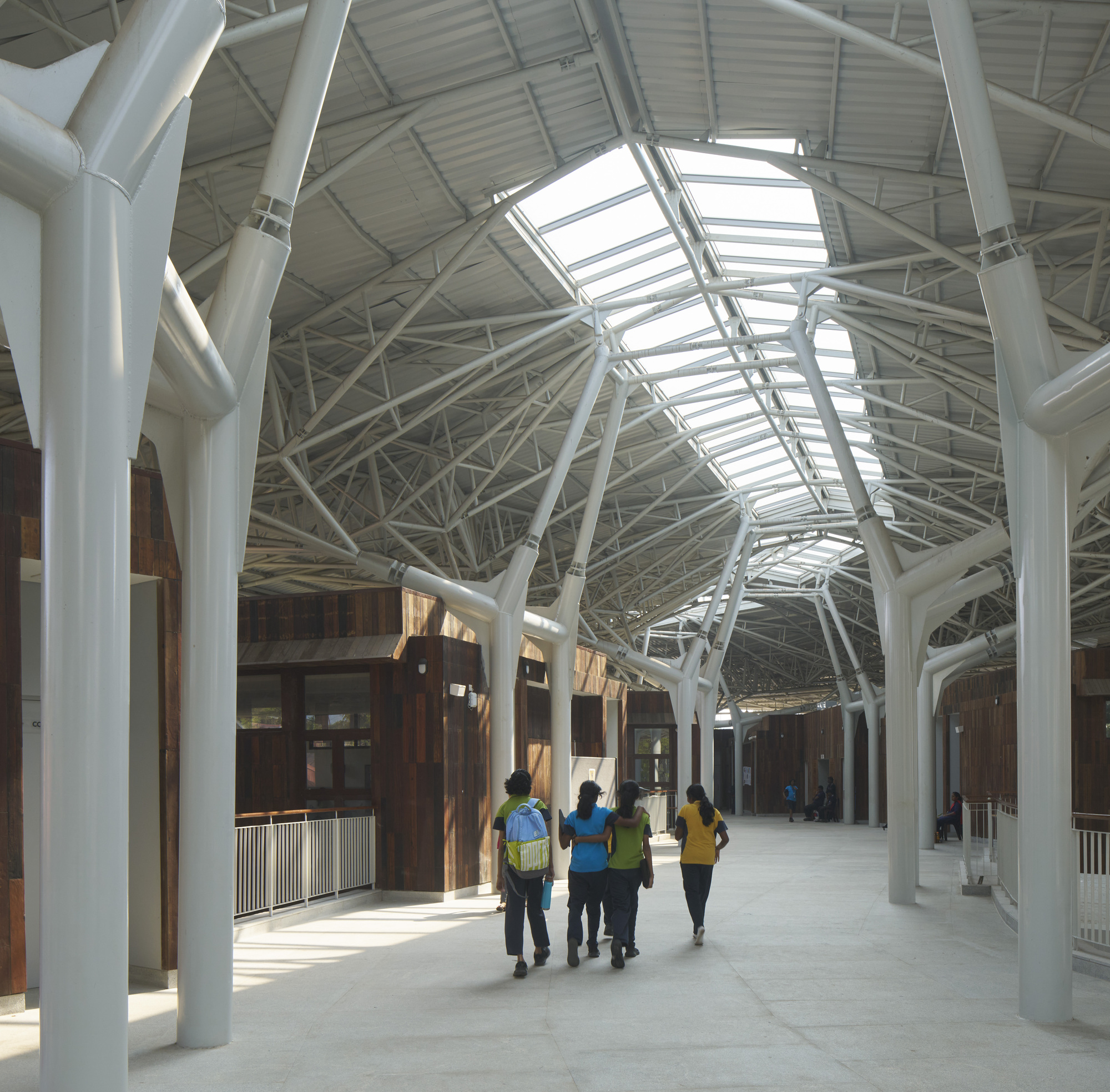 Interior circulation corridor with white structural trusses, skylights, and visitors walking past timber pavilions