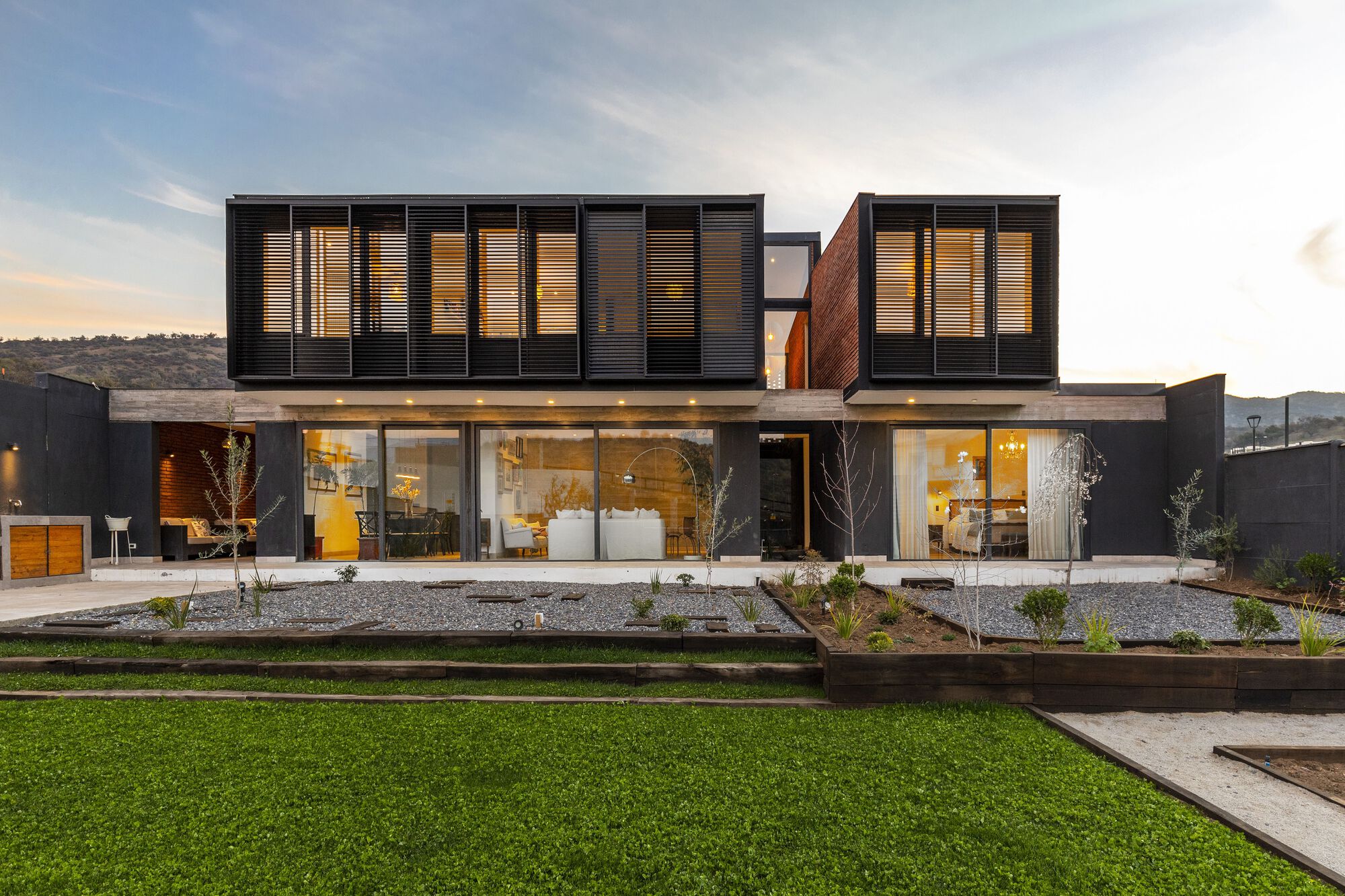 Garden facade with planted beds and floor-to-ceiling glazing beneath slatted black upper volume at twilight