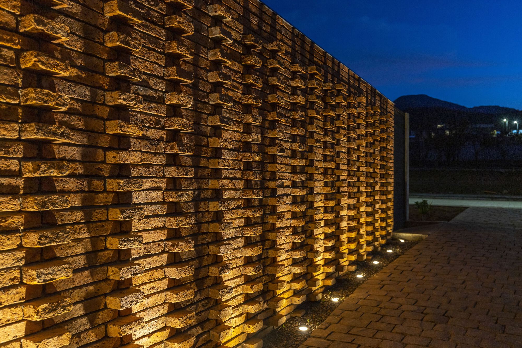 Close-up of staggered brick wall with ground-level uplighting illuminating the relief pattern at dusk